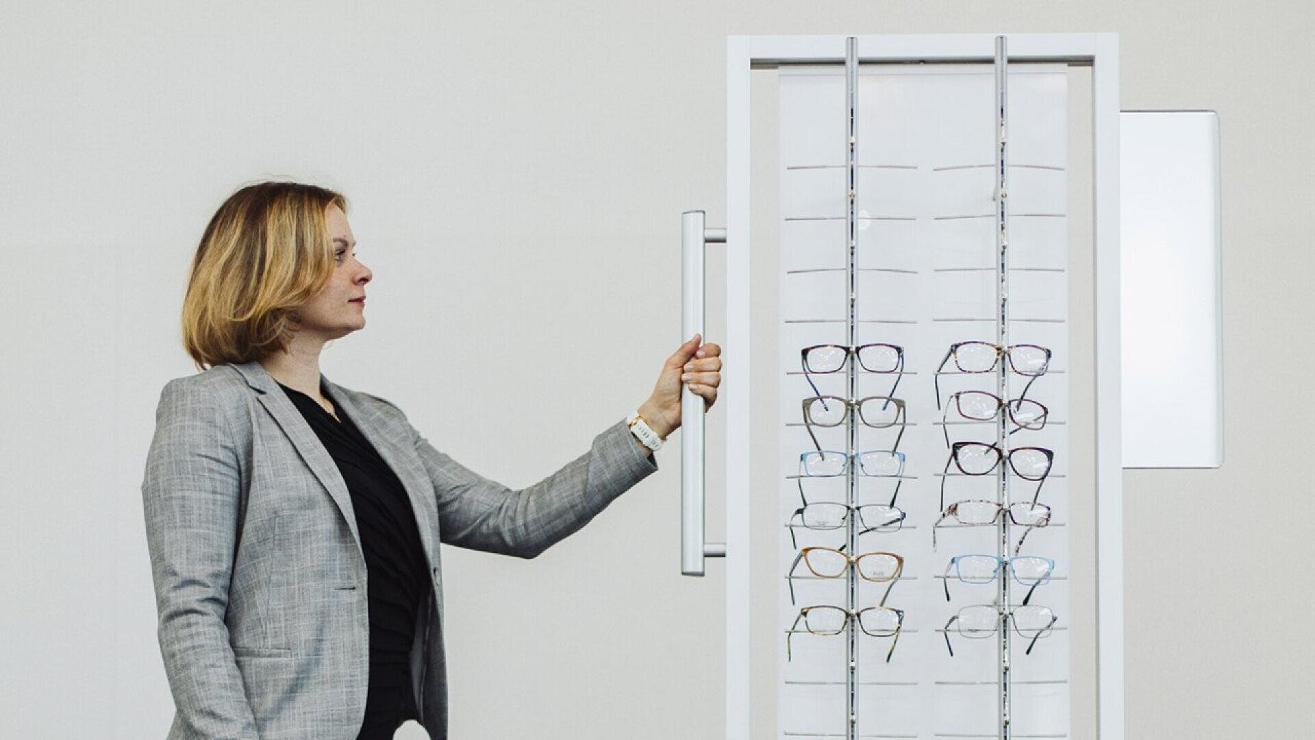 Eine Frau in einem grauen Blazer betrachtet ein Brillengestell mit mehreren Brillen vor einem schlichten, hellen Hintergrund.
