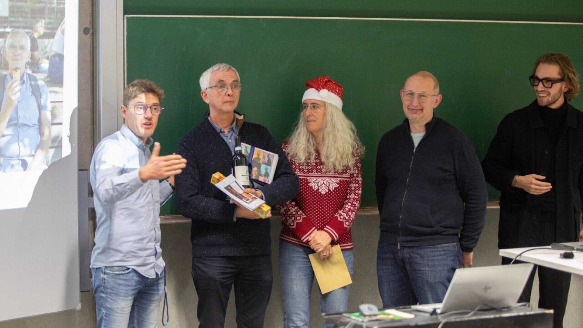 Fünf Erwachsene stehen vor einem Klassenzimmer. Einer gestikuliert beim Sprechen. Einer hält ein Geschenk in der Hand, ein anderer trägt eine Weihnachtsmannmütze und einen roten Pullover. Im Hintergrund sind eine Tafel und ein projiziertes Foto zu sehen.