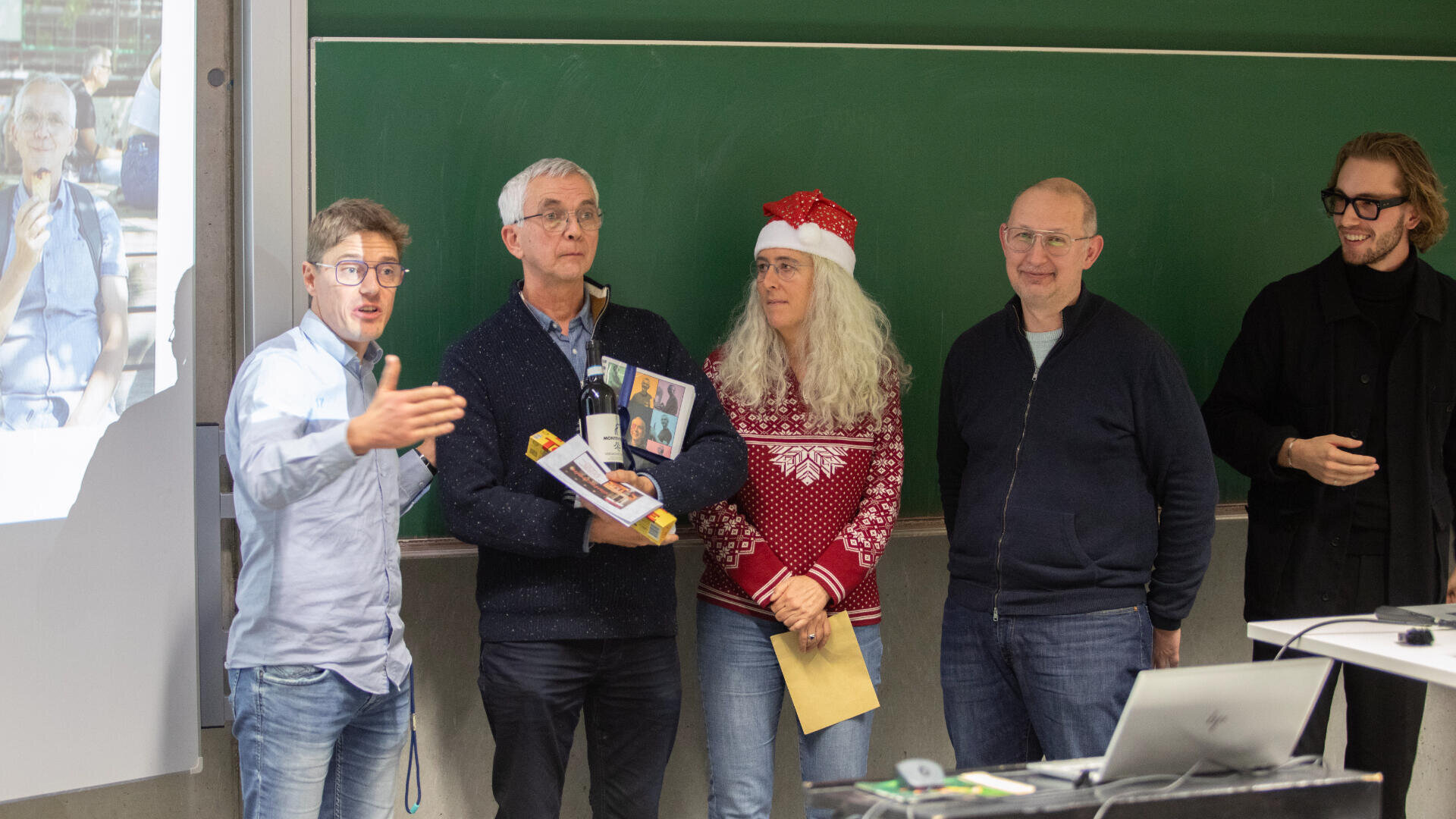 Five adults stand at the front of a classroom near a green chalkboard. One gestures while speaking, another holds a gift, and one woman wears a red Christmas sweater and a Santa hat. A projector screen is visible to the side.