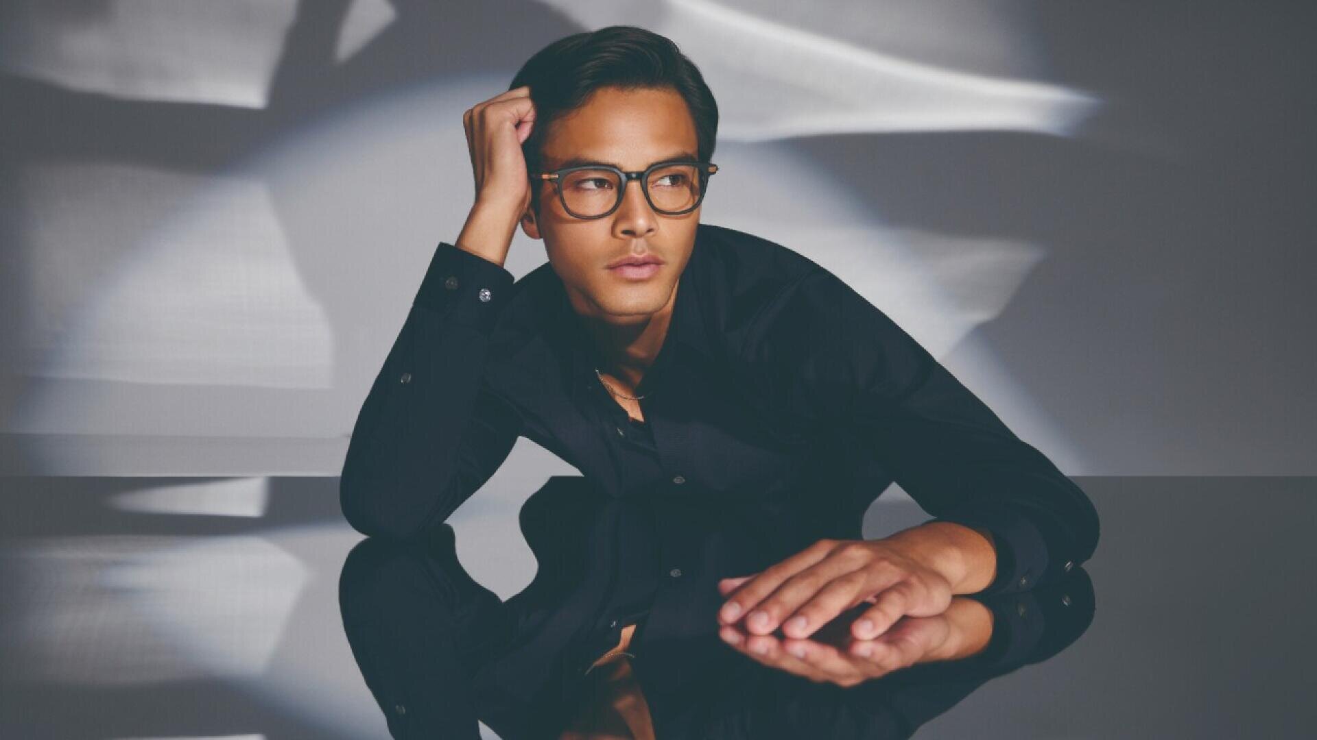 A man wearing glasses and a black shirt leans on a reflective surface, his head resting on one hand. Soft light and shadows create an artistic background.