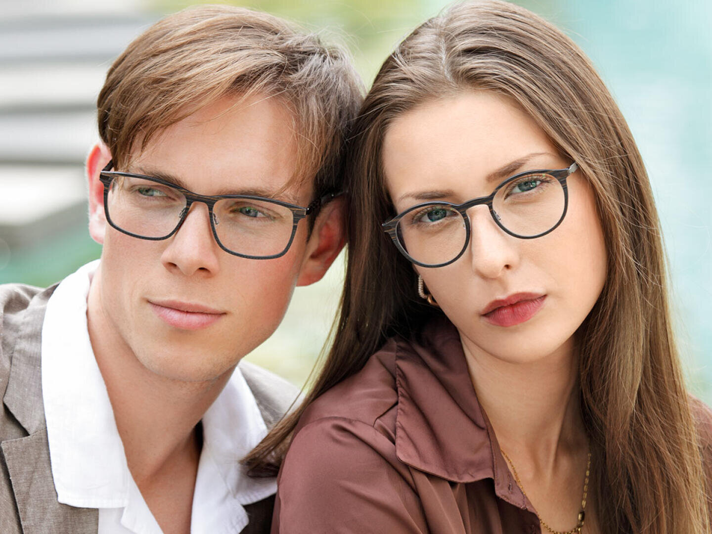 A young man and woman wearing glasses are sitting close together outdoors, looking slightly away from the camera. Both have light skin and straight brown hair. The background is blurred.