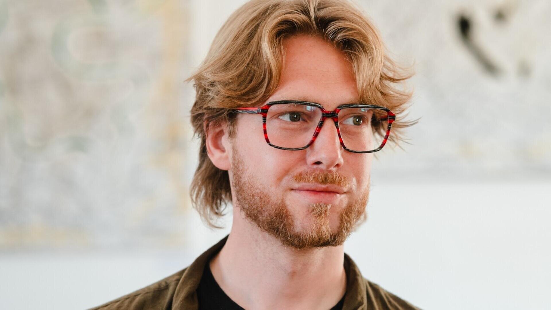 A man with light brown hair, a short beard, and mustache, wearing large, red-patterned glasses and a dark shirt, looks slightly to the side with a neutral expression. The background is softly blurred.