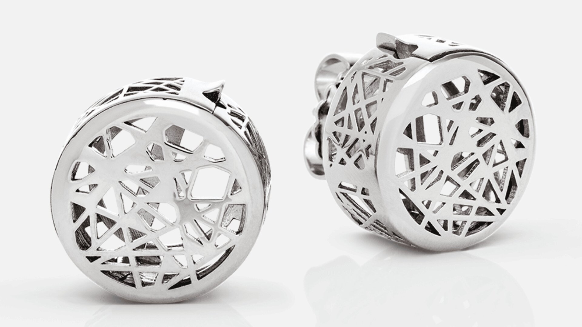 Ein Paar runde Silberohrringe mit geometrischen Ausschnittmustern auf weißem Untergrund. Das filigrane Design zeichnet sich durch sich kreuzende Linien und eckige Formen aus und verleiht ihnen ein modernes und elegantes Aussehen.