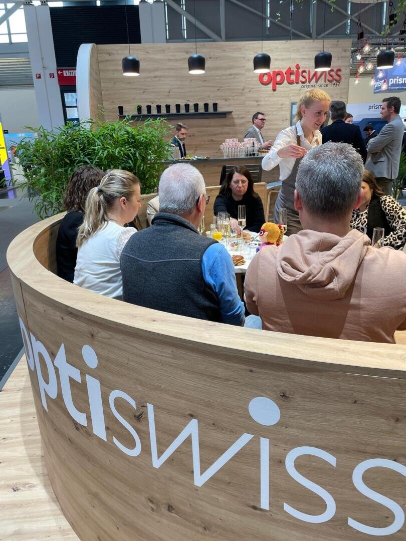 Eine Gruppe von Personen sitzt bei einer Veranstaltung um einen runden Holzstand herum, auf dem Getränke und Speisen stehen. An der Seite des Standes ist das optiswiss-Logo angebracht. Ein Mann steht und spricht zu der Gruppe. Moderne Dekoration und Pflanzen sind zu sehen.