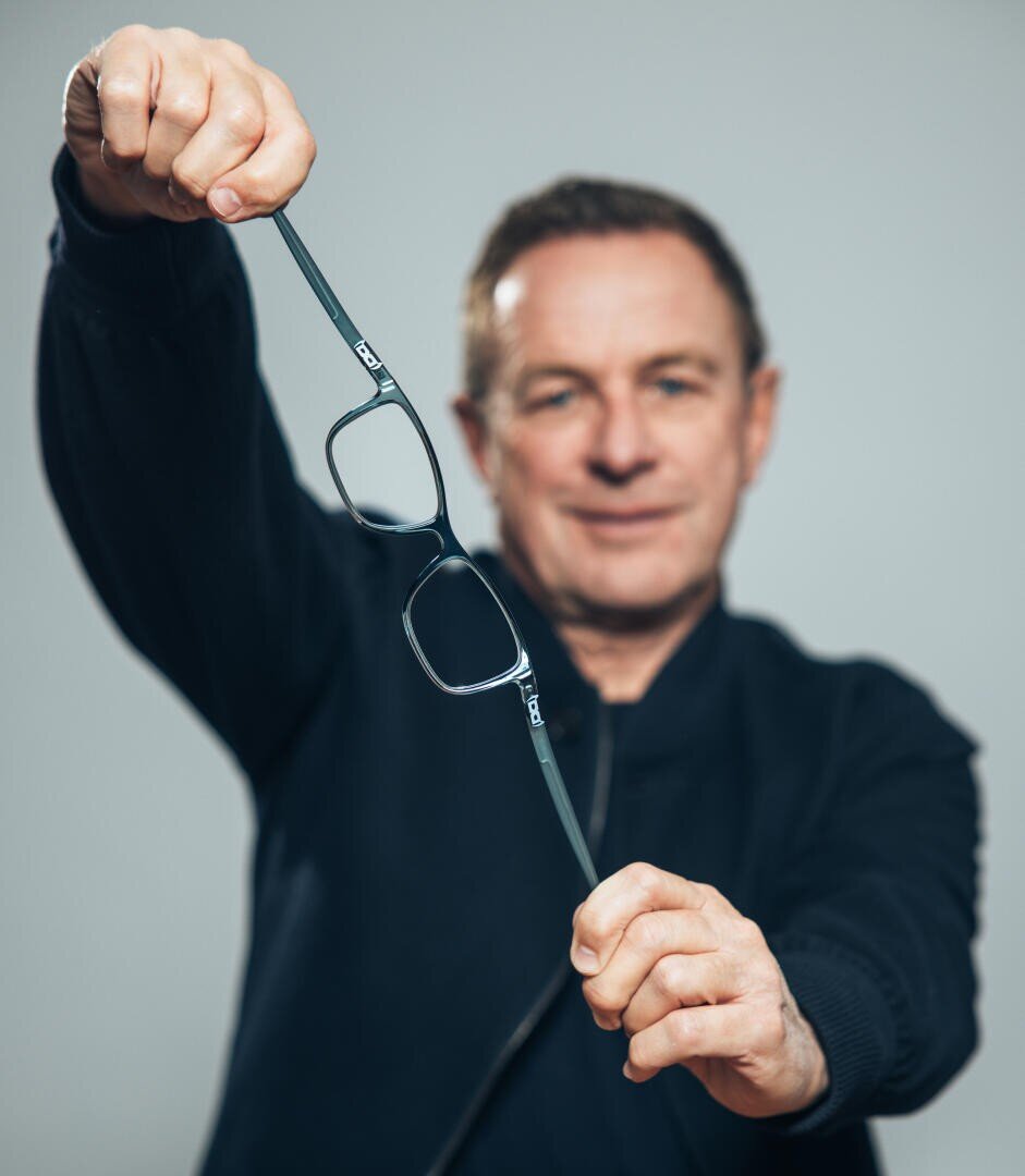 Ein Mann in einer dunklen Jacke hält mit beiden Händen eine Brille in Richtung der Kamera und fokussiert auf die Brille, während sein Gesicht im Hintergrund unscharf erscheint.