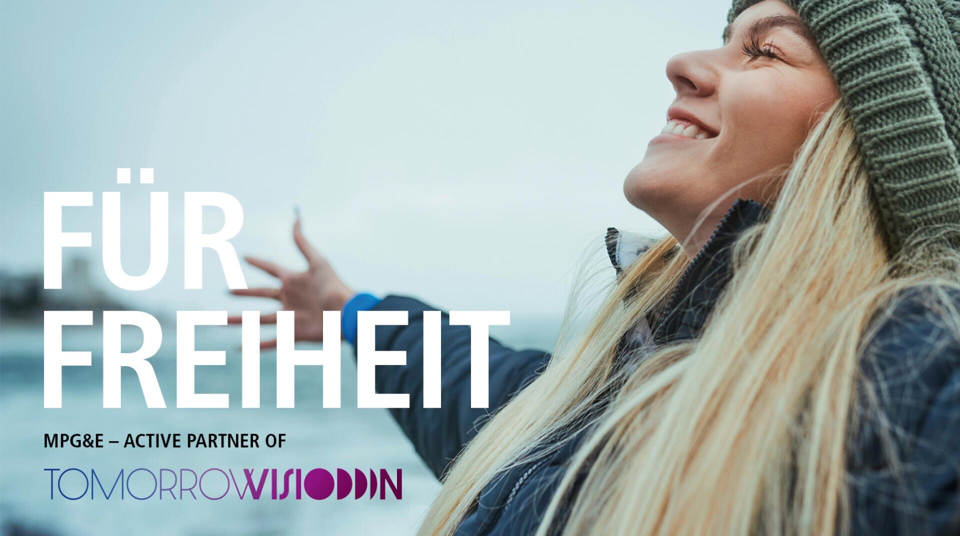 Eine lächelnde Frau mit langen blonden Haaren steht im Freien am Wasser, die Arme ausgestreckt, und genießt die frische Luft. Der große Text lautet "FÜR FREIHEIT". Am unteren Rand erscheinen die Logos von MPG&E und Tomorrow Wisdom.