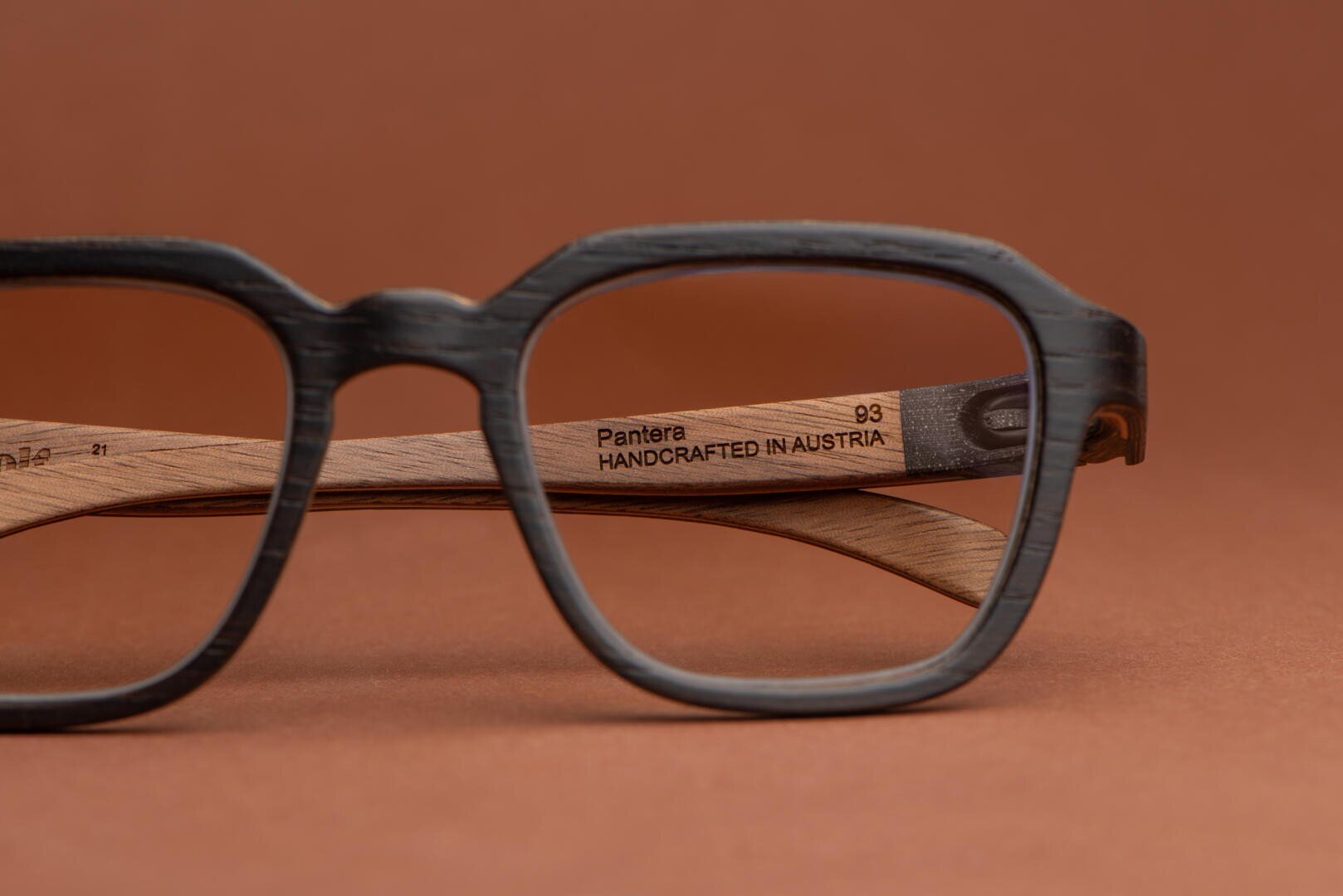 Close-up of stylish eyeglass frames with black rims and wooden arms, showing the engraved text Pantera HANDCRAFTED IN AUSTRIA on the inner side. The background is a matching brown color.