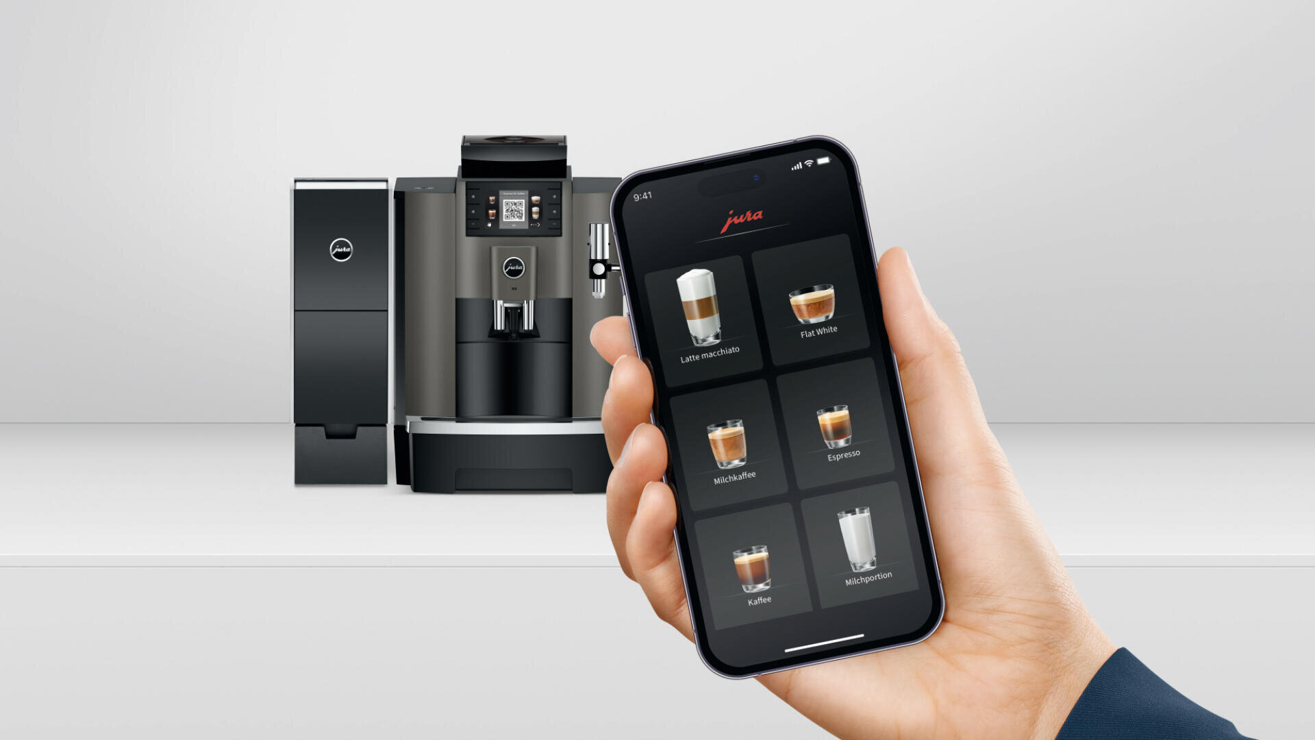 Eine Hand hält ein Smartphone, auf dem die App-Oberfläche einer Kaffeemaschine mit verschiedenen Kaffeeoptionen angezeigt wird. Im Hintergrund steht ein moderner Kaffeevollautomat auf einer weißen Fläche.