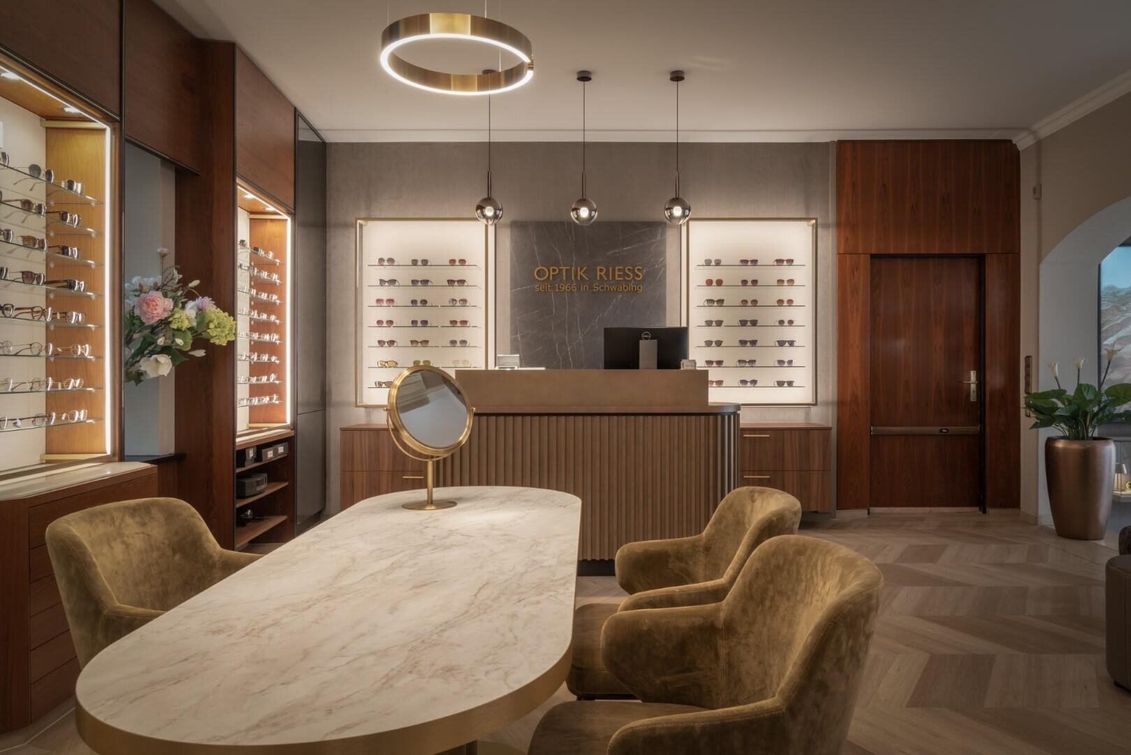 Ein stilvolles Optikergeschäft mit Marmortisch, Samtstühlen, Brillenregalen, Empfangstresen, Hängelampen, Holzakzenten und Blumenschmuck. Auf dem Schild steht „Optik Riess“.