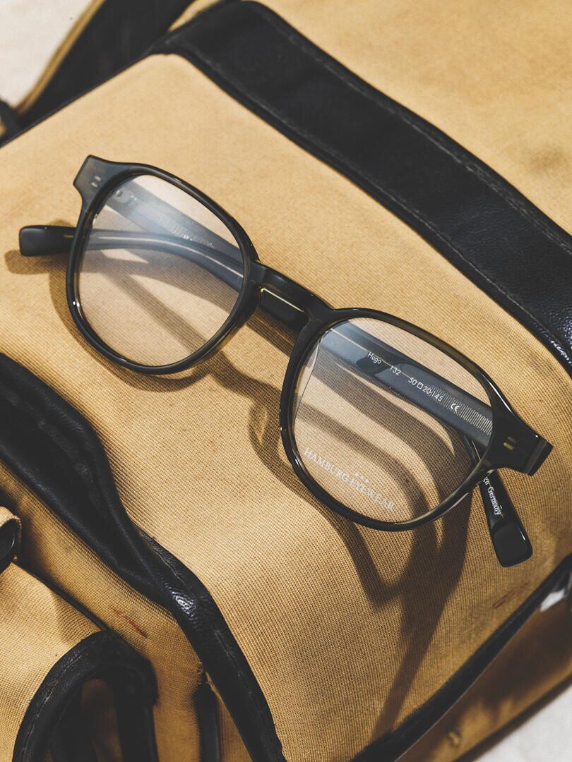 Eine schwarz umrandete Brille liegt auf einem braun-schwarzen Lederrucksack, in dem sich das Licht auf den Gläsern spiegelt.