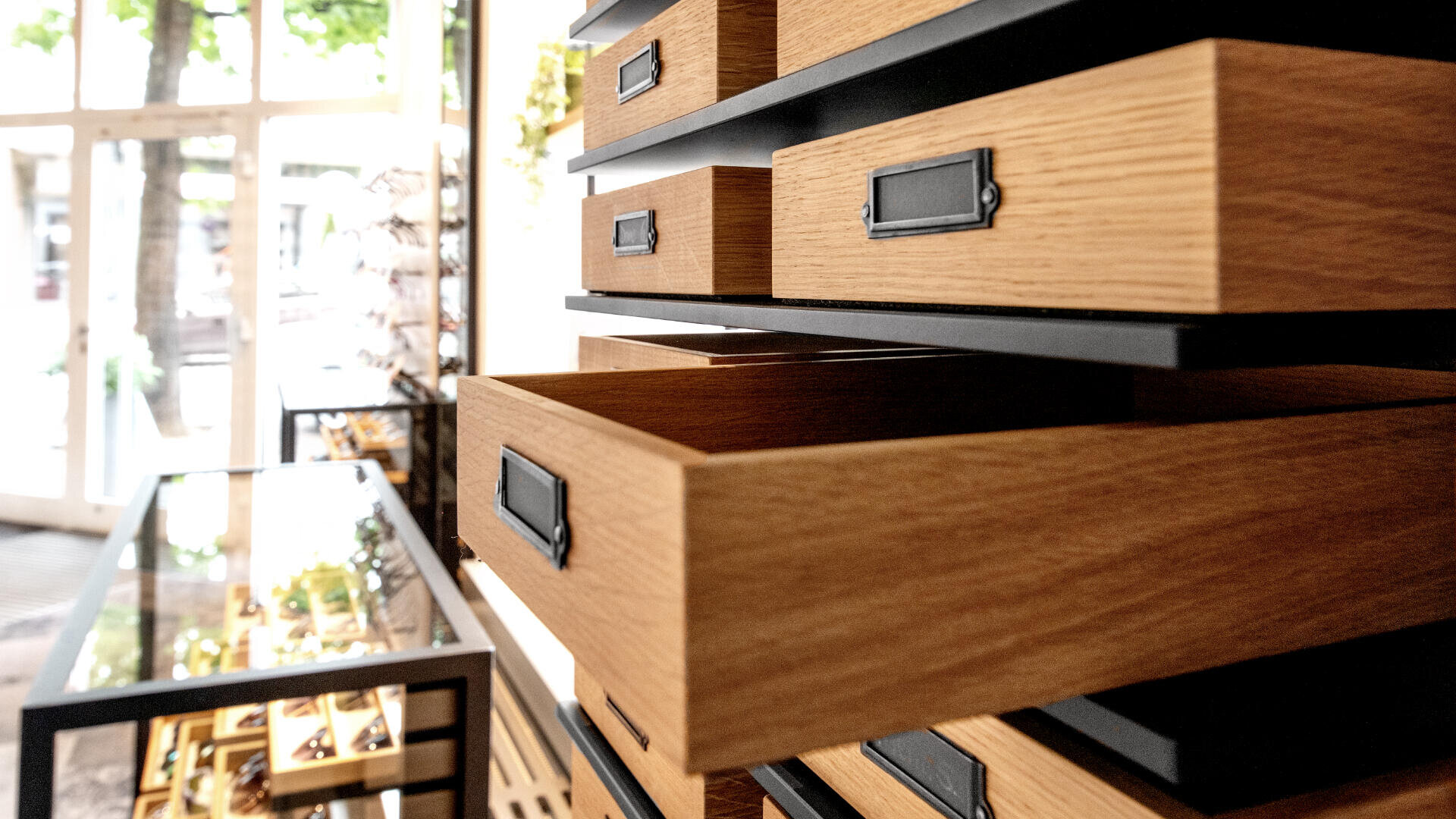 Nahaufnahme mehrerer Holzschubladen, einige davon teilweise geöffnet, vor einem modernen Interieur mit Glasvitrinen und großen Fenstern, die natürliches Licht hereinlassen.