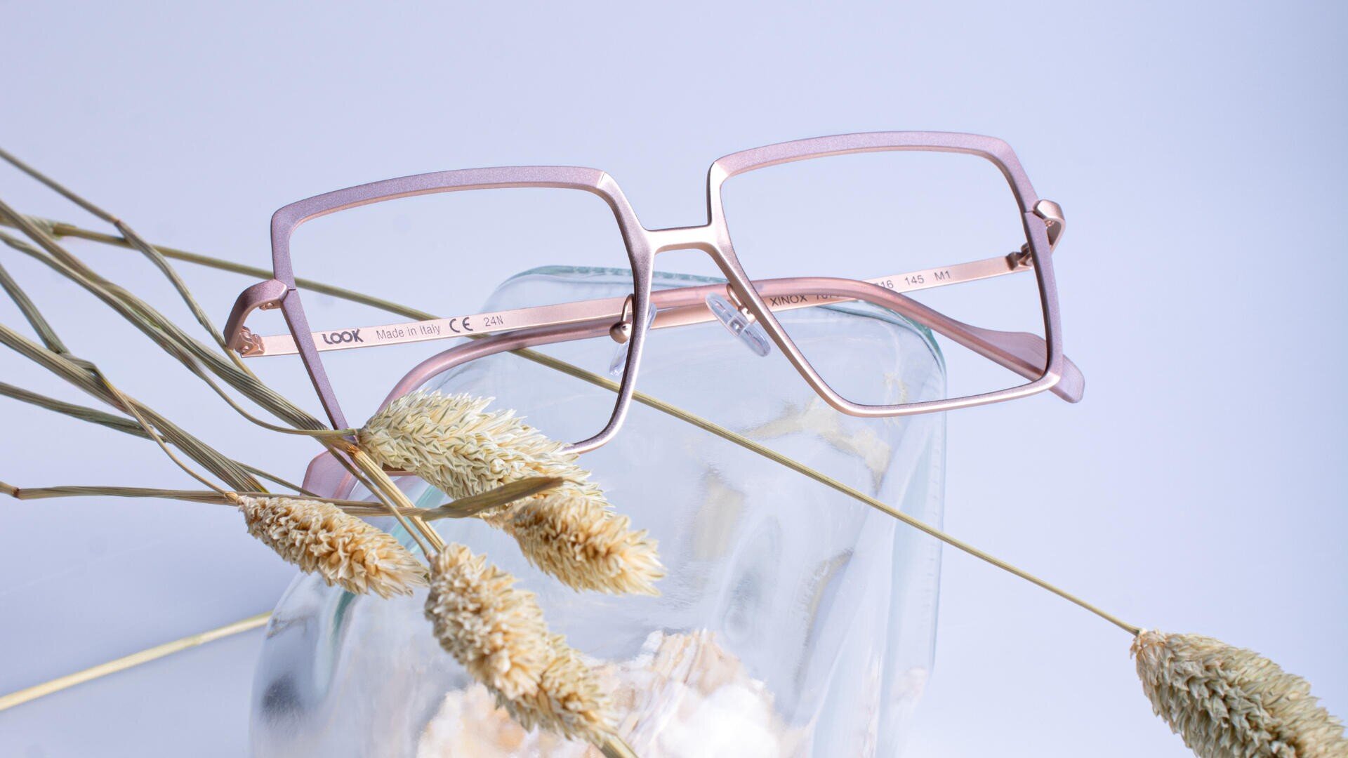 A pair of square, pink-rimmed eyeglasses rests on a clear glass jar, surrounded by dried beige plants against a pale blue background.