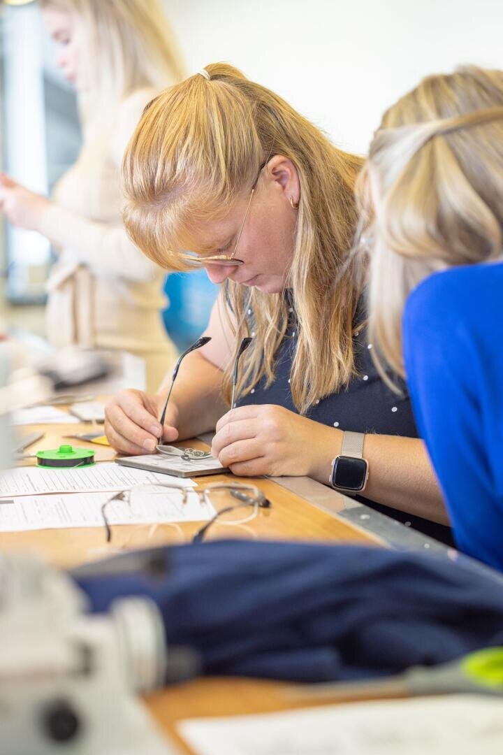 Eine Frau mit langen blonden Haaren und Brille arbeitet konzentriert mit kleinen Werkzeugen an einem Schreibtisch, umgeben von Papieren und Geräten, während eine andere Person in Blau neben ihr sitzt.