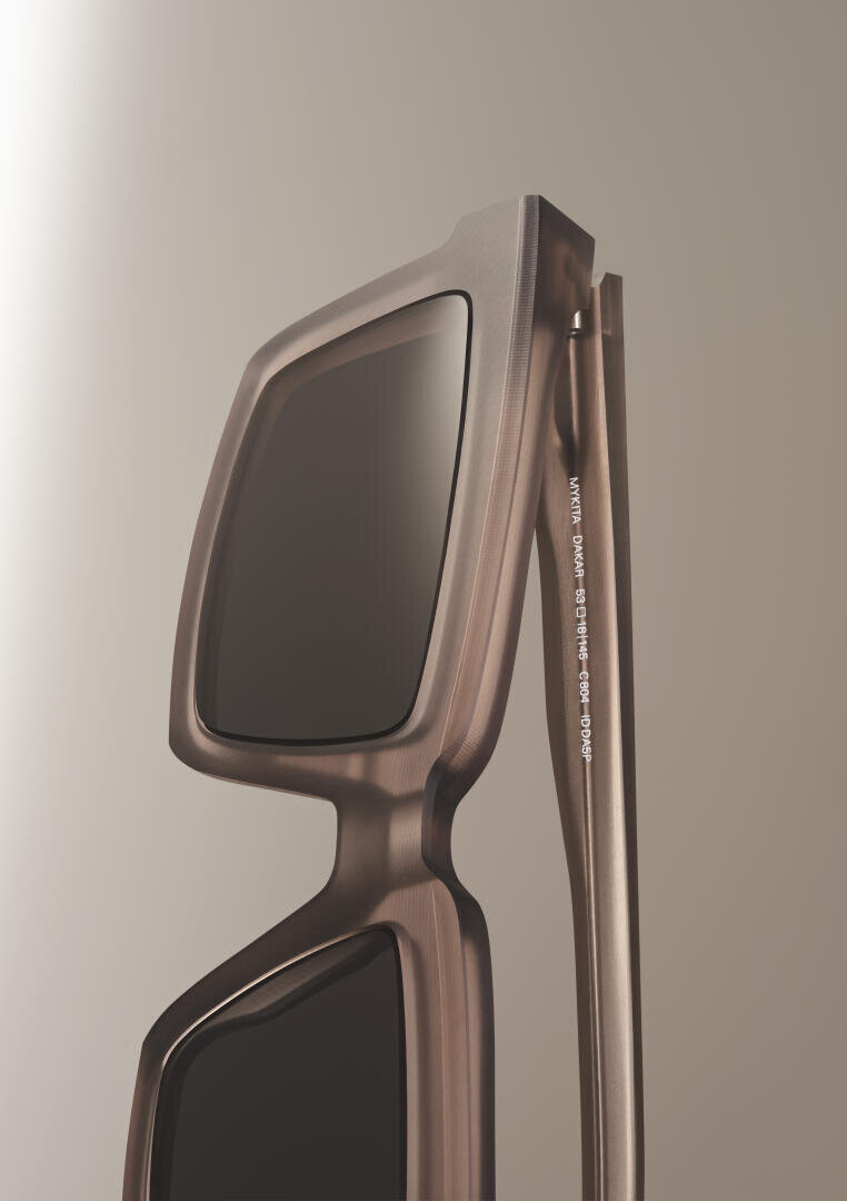 Close-up view of a pair of modern, square-shaped sunglasses with thick, metallic frames and dark lenses, positioned against a neutral beige background.