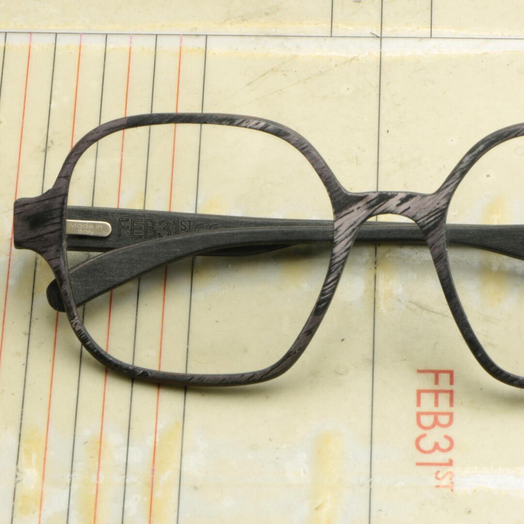 Eine schwarze, quadratisch gerahmte Brille liegt auf einem vergilbten Papierformular mit rotem Text, auf dem FEB 31st steht. Die Brille ist so positioniert, dass der linke Bügel nach innen geklappt ist.