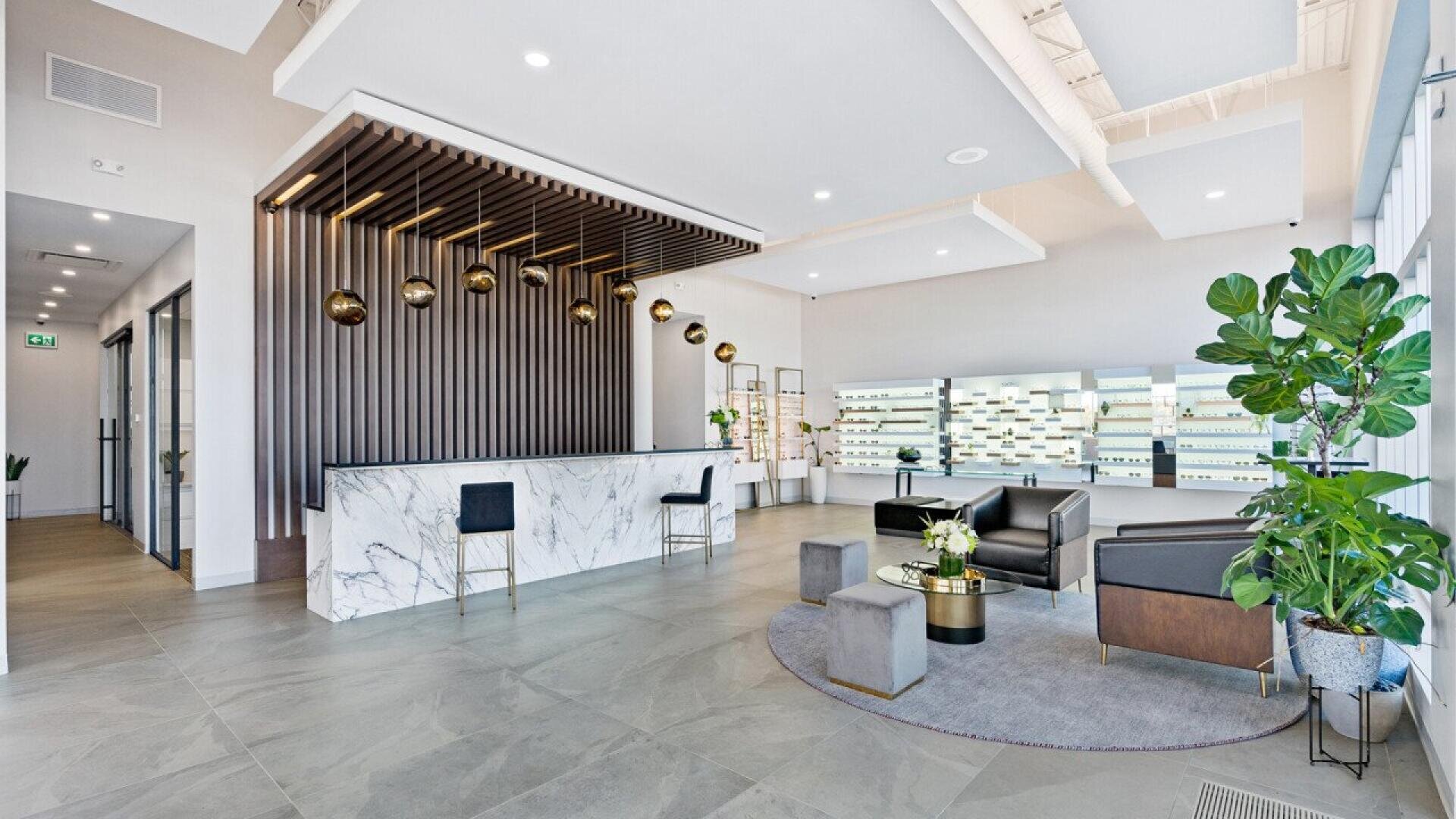 Modern office lobby with marble reception desk, gold hanging lights, black chairs, gray tile floor, and large windows. There are plants, a seating area with black armchairs, and a wall display in the background.