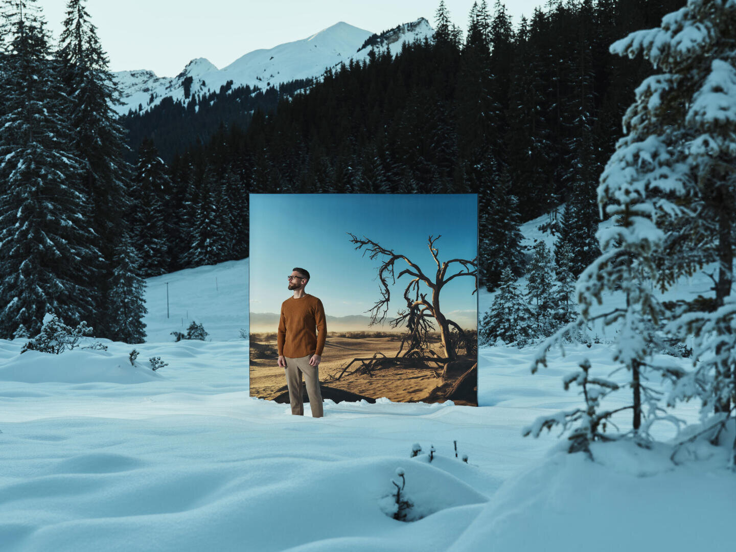 A man stands in the deep snow between pine trees and mountains. Behind him is a large photo of a dry, barren landscape with a dead tree that forms a stark contrast to the snow-covered surroundings.