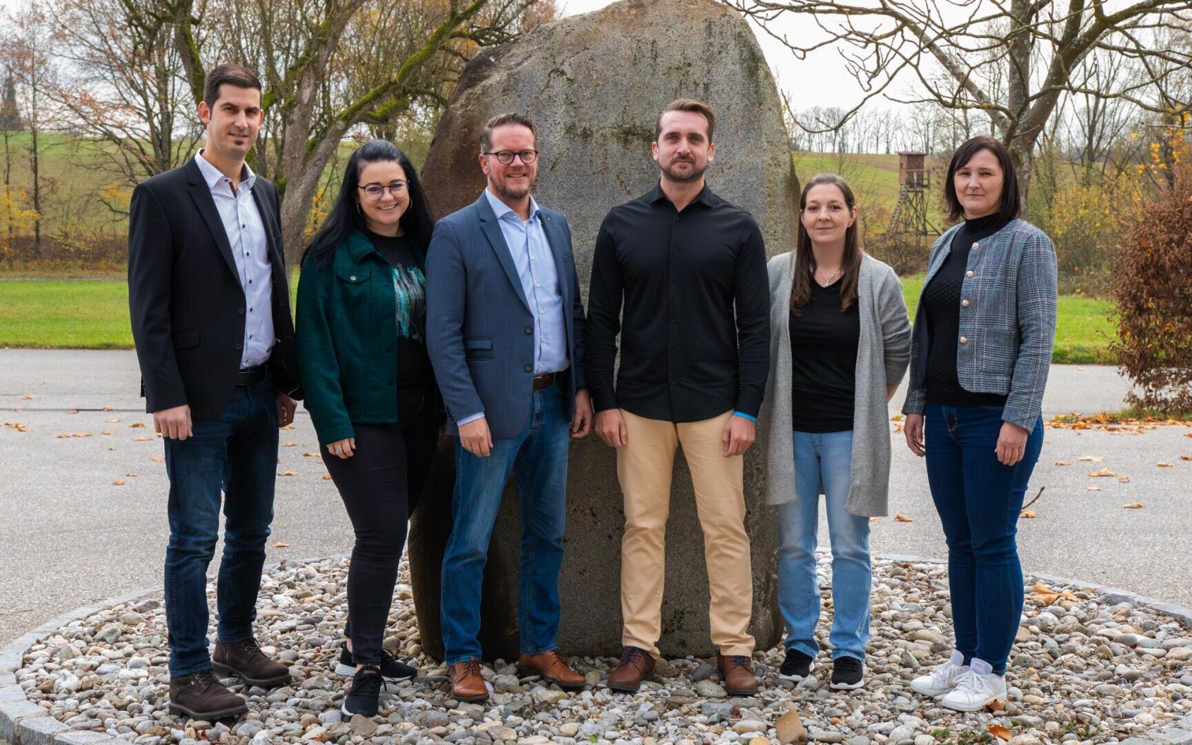 Sechs Erwachsene stehen im Freien vor einem großen Felsen auf einer mit Kieselsteinen bedeckten Fläche, mit Bäumen und Gras im Hintergrund. Sie sind in Freizeit- und Business-Casual-Kleidung gekleidet und posieren mit dem Gesicht zur Kamera für ein Gruppenfoto.