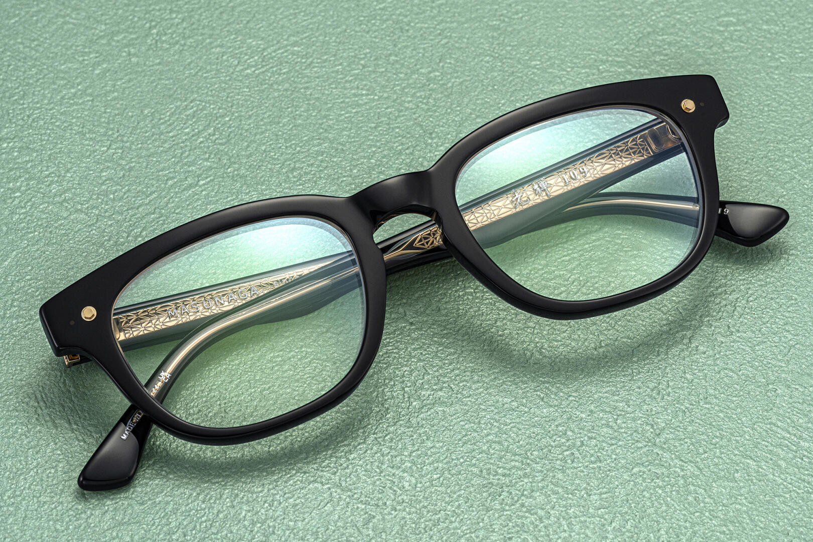 Eine schwarz umrandete Brille mit goldenen Verzierungen an den Bügeln, die auf einer grün strukturierten Oberfläche platziert ist.