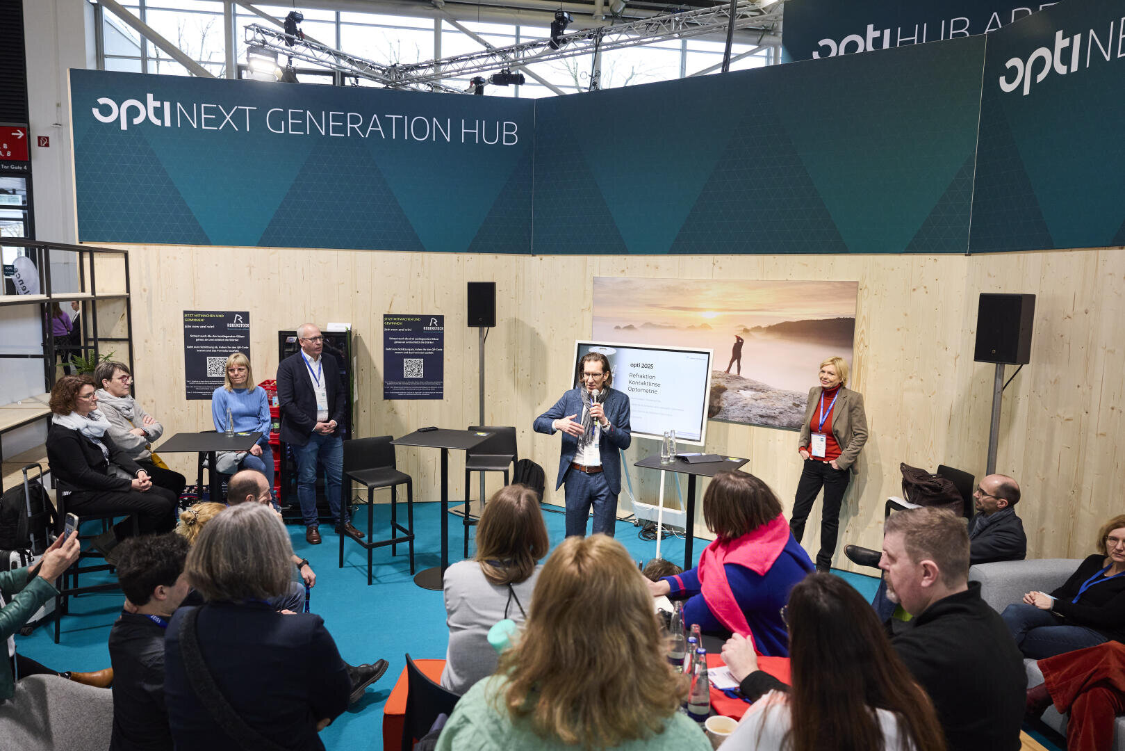 Ein Redner mit einem Mikrofon spricht zu einem sitzenden Publikum bei einer Indoor-Veranstaltung, in dessen Nähe mehrere Personen stehen. Auf dem Bühnenhintergrund steht opti NEXT GENERATION HUB. Die Umgebung wirkt modern und professionell.