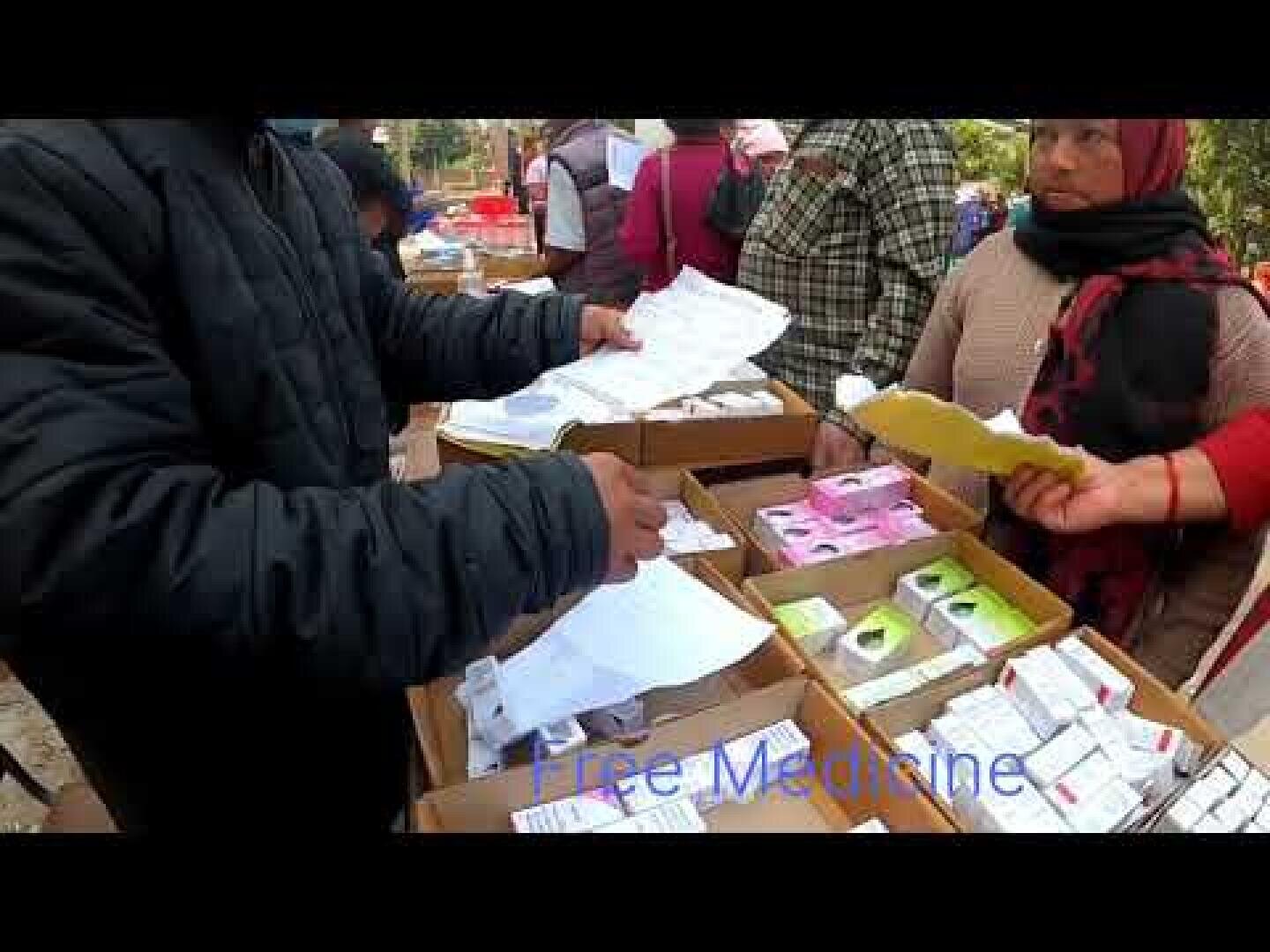 Eine Person verteilt Papiere in der Nähe von Schachteln mit verschiedenen Medikamenten an einem Stand im Freien, während andere in der Nähe stehen. Der Schriftzug Free Medicine ist am unteren Rand des Bildes schwach zu erkennen.