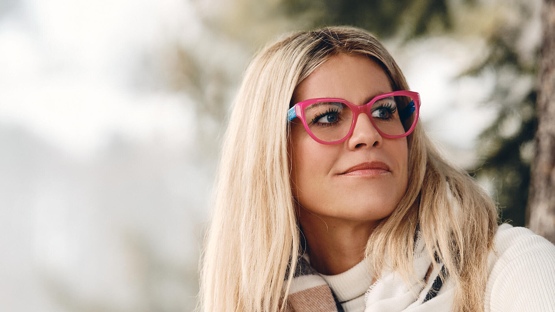 Eine Frau mit langen blonden Haaren, einer leuchtend rosa Brille und einem hellen Pullover blickt nachdenklich zur Seite ins Freie, im Hintergrund sind verschwommene Bäume zu sehen.