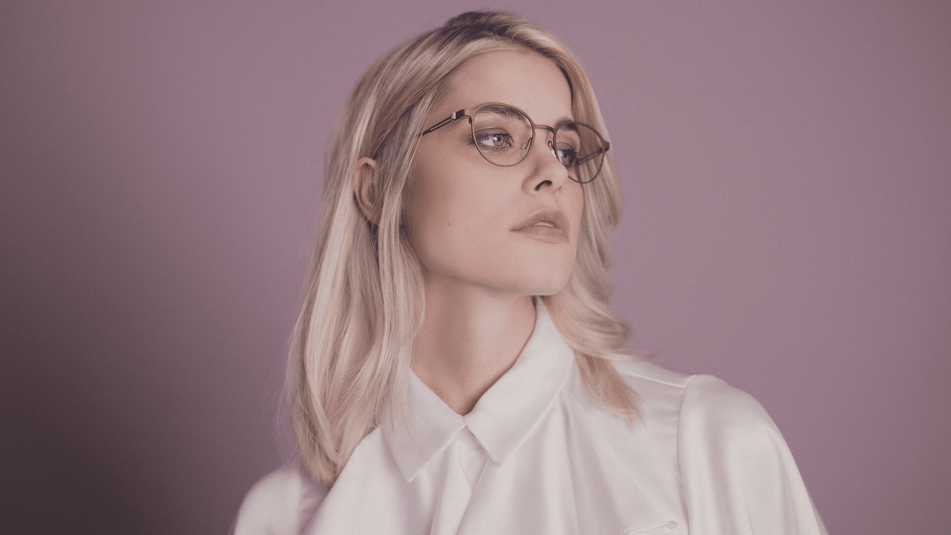 Eine Frau mit schulterlangem blondem Haar, Brille und weißem Hemd mit Kragen blickt vor einem gedämpften violetten Hintergrund zur Seite.