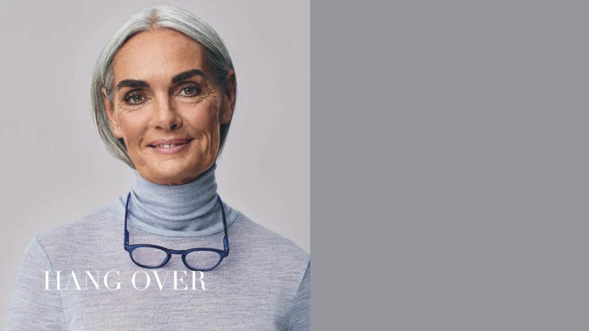 A smiling older woman with gray hair and blue eyes wears a light gray turtleneck and blue glasses around her neck. The text HANG OVER appears in white on the left. The background is light gray.