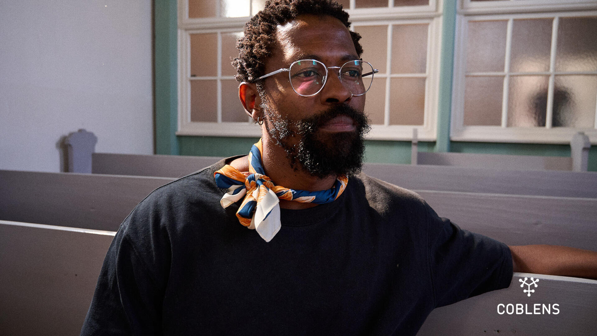 A man with glasses, a beard, and a colorful scarf sits on a light gray church pew in a sunlit room. He gazes thoughtfully into the distance. The Coblens logo is visible in the bottom right corner.
