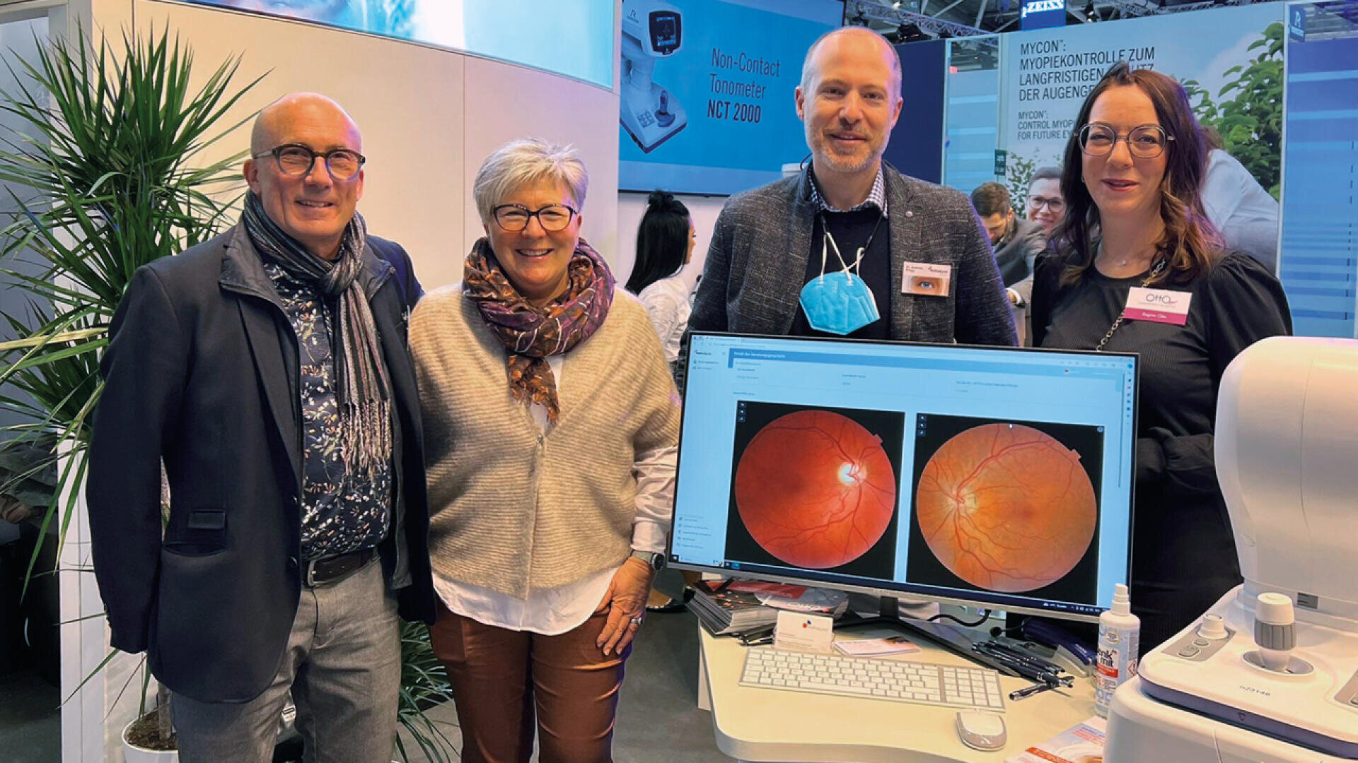 Vier Erwachsene stehen lächelnd an einem Messestand mit medizinischem Gerät. Ein Computerbildschirm zeigt Netzhautbilder. Im Hintergrund sind eine Topfpflanze und Informationsplakate zu sehen.