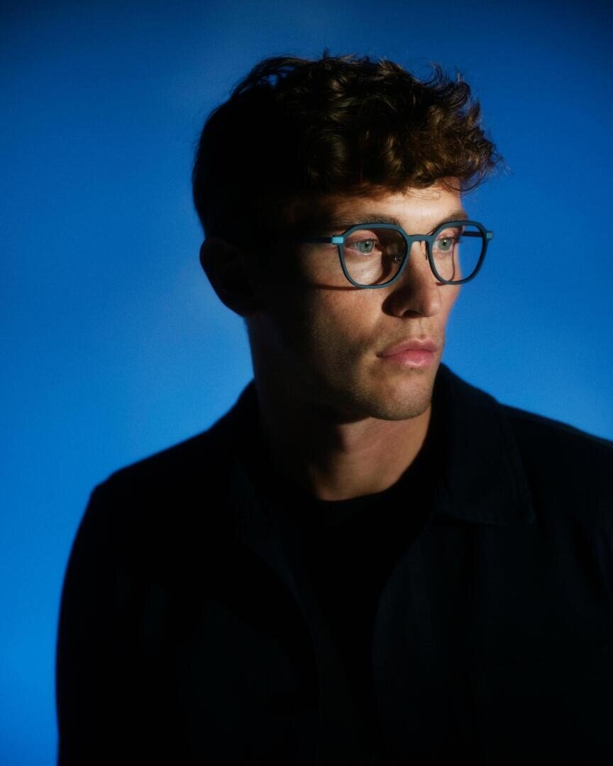 A young man with curly brown hair and glasses looks to the side against a blue background, with dramatic lighting creating shadows on his face and clothing.