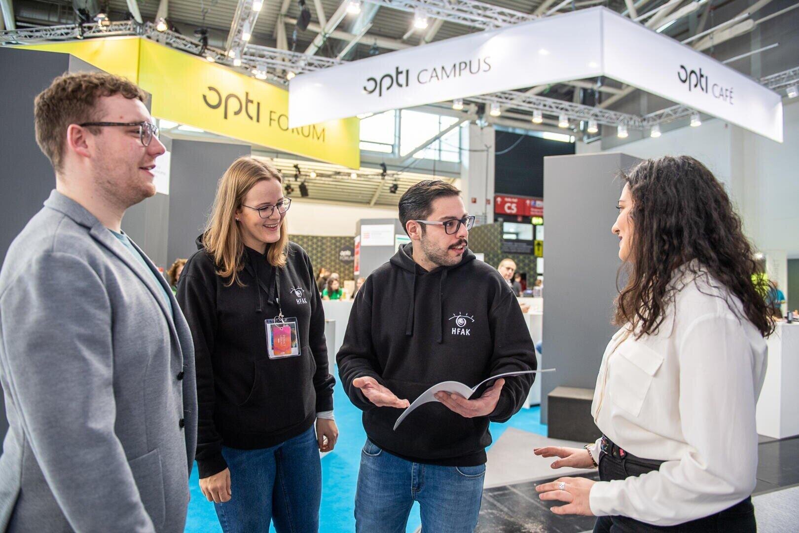 Vier Personen unterhalten sich auf einer Indoor-Veranstaltung oder -Messe, wobei zwei von ihnen schwarze Kapuzenpullis tragen und einer eine aufgeschlagene Broschüre in der Hand hält. Sie lächeln und scheinen in ein Gespräch vertieft zu sein, während im Hintergrund die Stände der Veranstaltung zu sehen sind.