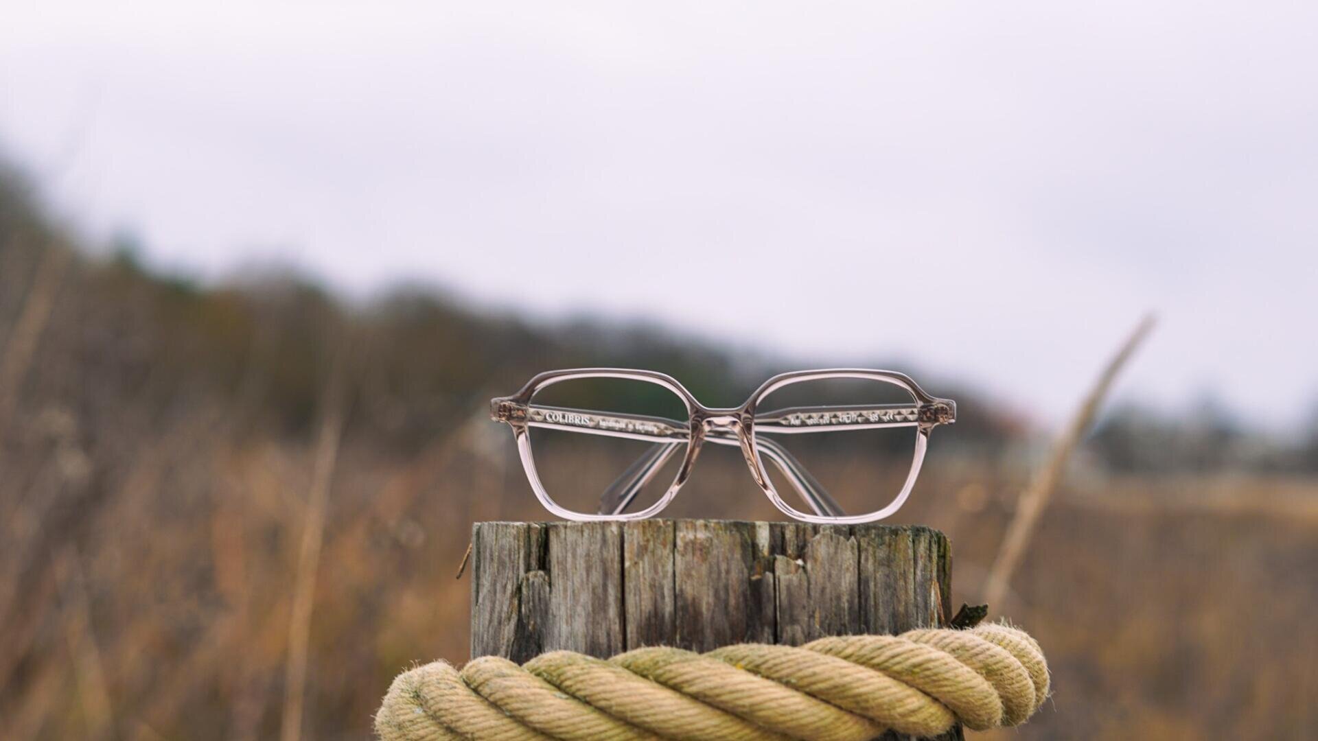 Eine Brille ruht auf einem verwitterten Holzpfosten, um den ein dickes Seil gewickelt ist; der Hintergrund ist unscharf und zeigt eine natürliche Umgebung.