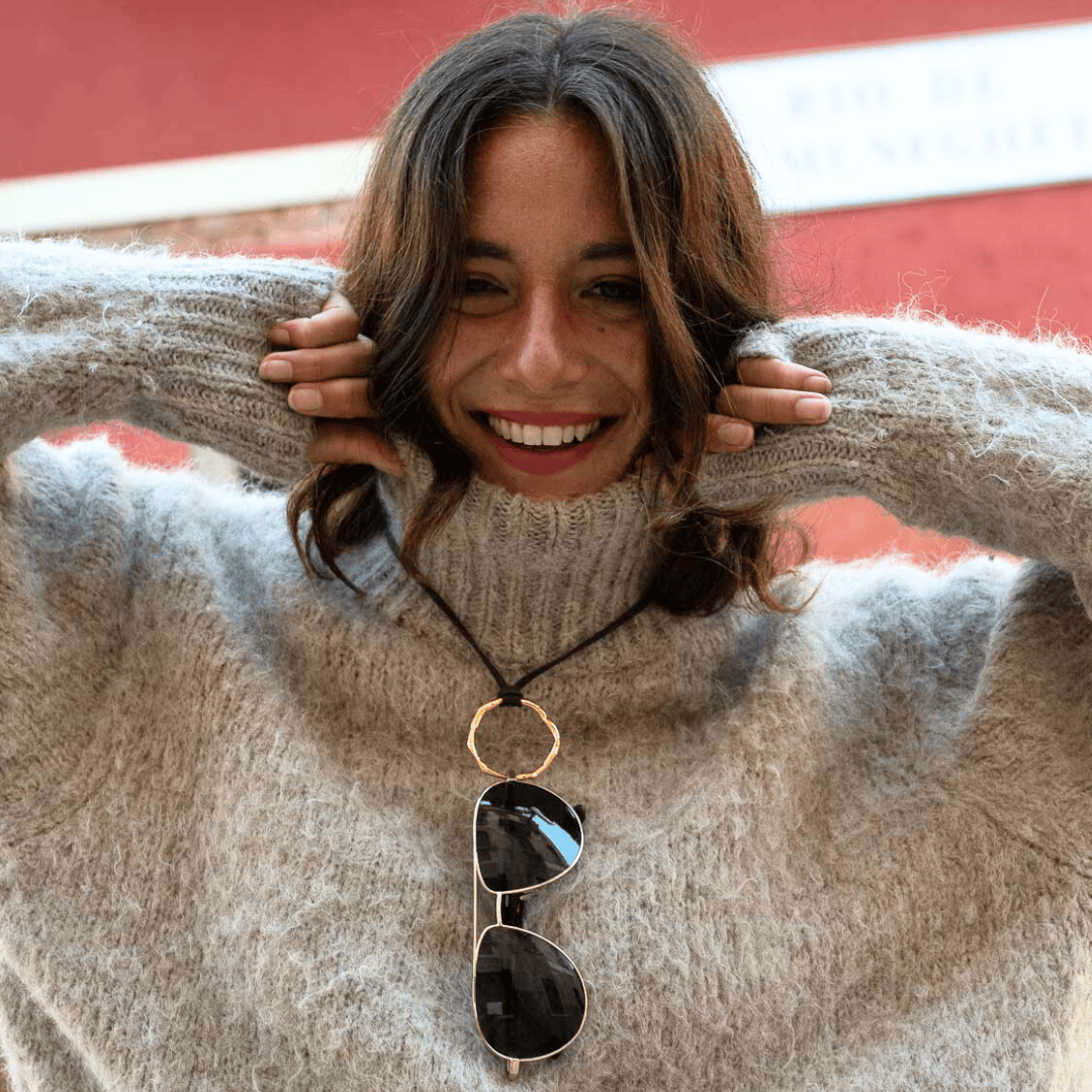 A smiling woman in a fuzzy beige sweater pulls the collar up to her chin, holding it with both hands. Sunglasses hang from a circular pendant necklace around her neck. A blurred red wall is in the background.