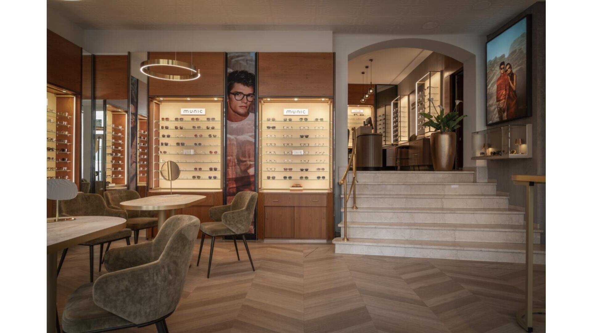 A stylish eyewear store interior with wooden floors, modern chairs, display cases filled with glasses, and warm lighting. A staircase leads to an upper level, and large eyewear ads decorate the walls.