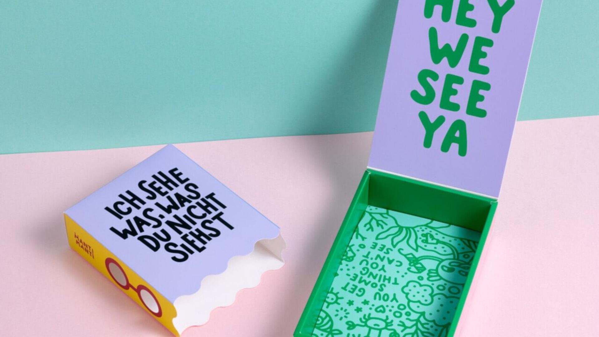 An open, colorful box with playful text; the lid says HEY WE SEE YA in green, and the side of the inner box reads ICH SEHE WAS, WAS DU NICHT SIEHST in black on purple. The background is pastel pink and green.