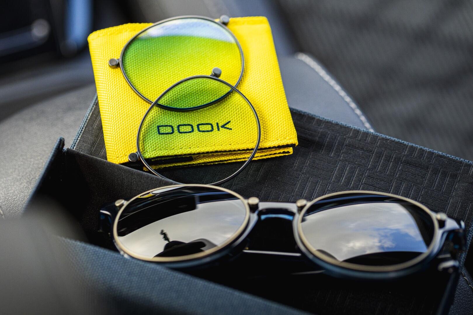 Eine runde schwarze Sonnenbrille mit verspiegelten Gläsern liegt in einer schwarzen Schachtel, während ein weiteres Glas und ein gelb-grünes Reinigungstuch mit der Aufschrift BOOK daneben liegen.
