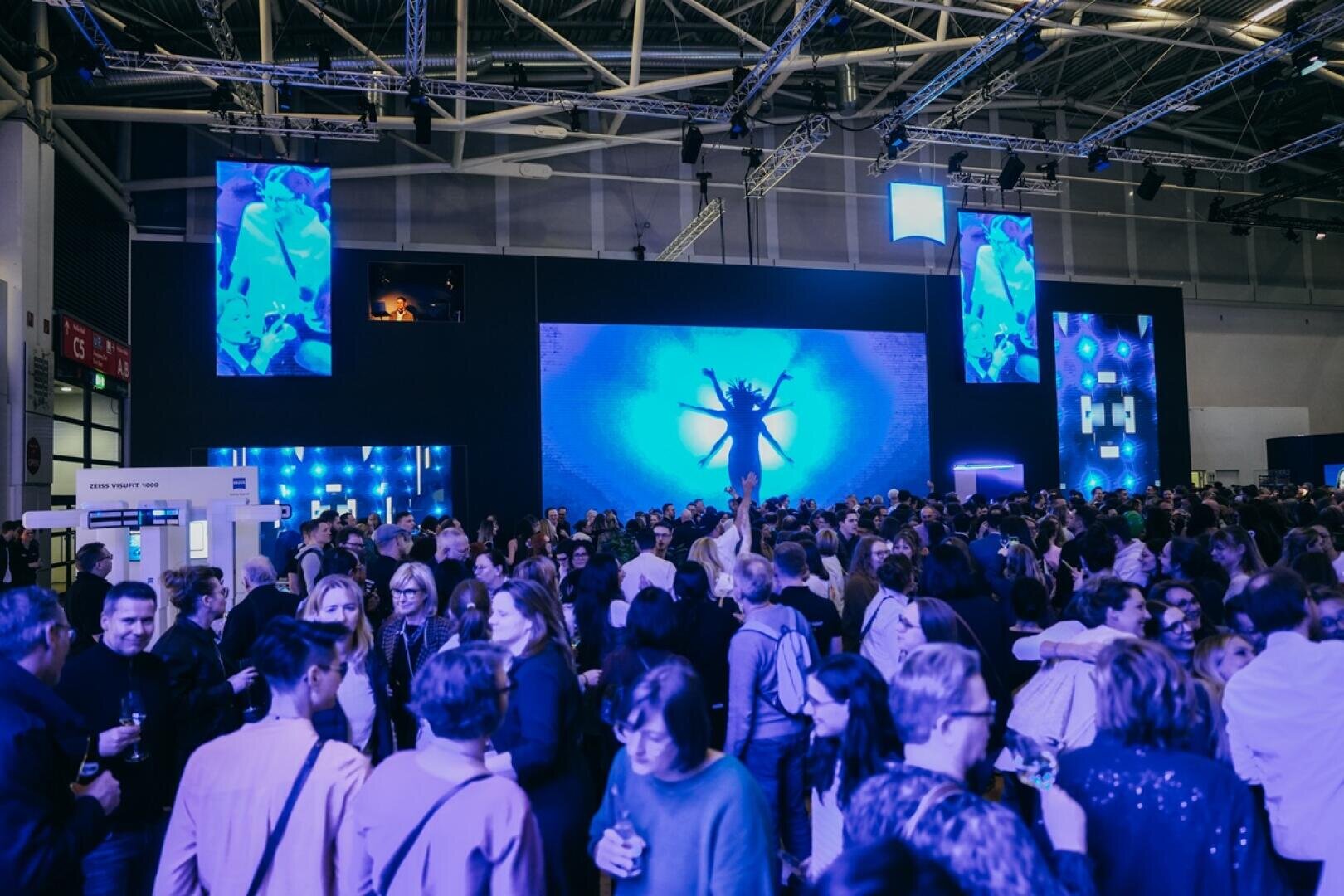 Eine große Menschenmenge versammelt sich bei einer Indoor-Veranstaltung mit Menschen, die sich unterhalten. Ein großer Bildschirm auf der Bühne zeigt blaue Grafiken, die ein Insekt darstellen. Oberlichter und kleinere Bildschirme tragen zu der lebhaften Atmosphäre bei.