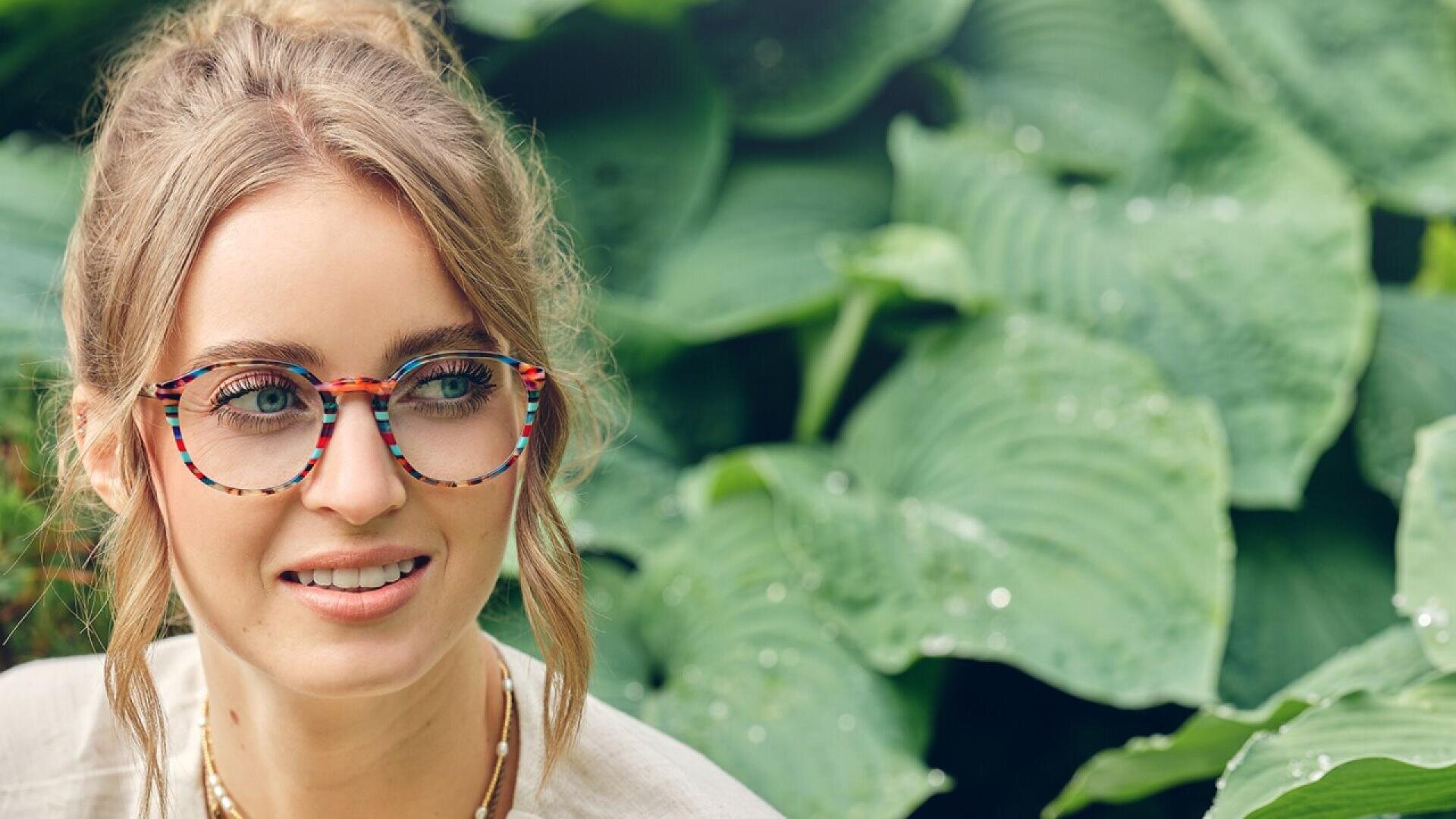 A woman with light brown hair in a loose bun, wearing colorful patterned glasses and layered necklaces, smiles while standing in front of large green leafy plants.