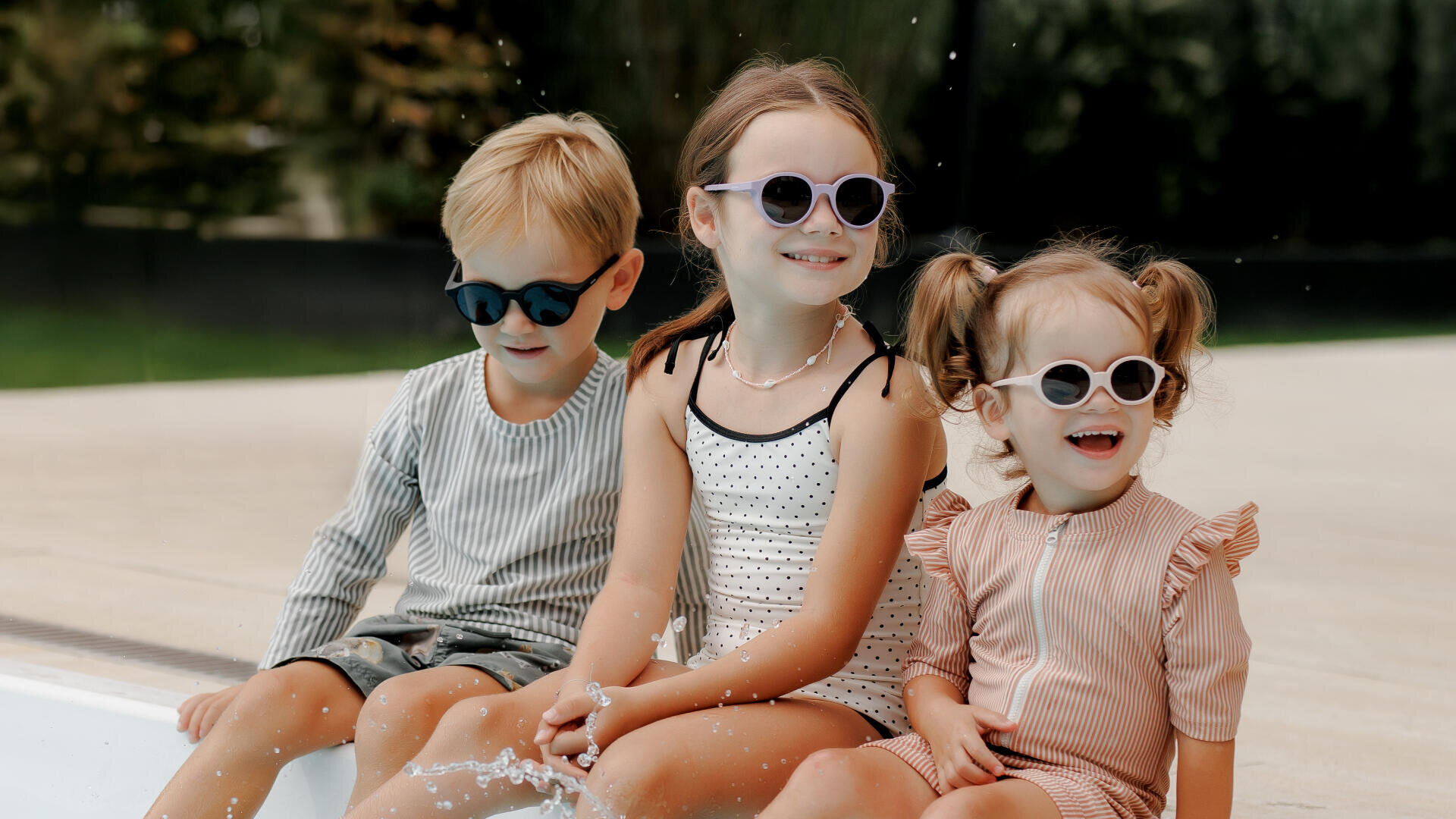 Drei kleine Kinder mit Sonnenbrillen sitzen lächelnd an einem Pool und planschen im Wasser. Der Junge trägt ein gestreiftes Outfit, das Mädchen in der Mitte einen gepunkteten Badeanzug und das Mädchen rechts ein pfirsichfarbenes Kleid.