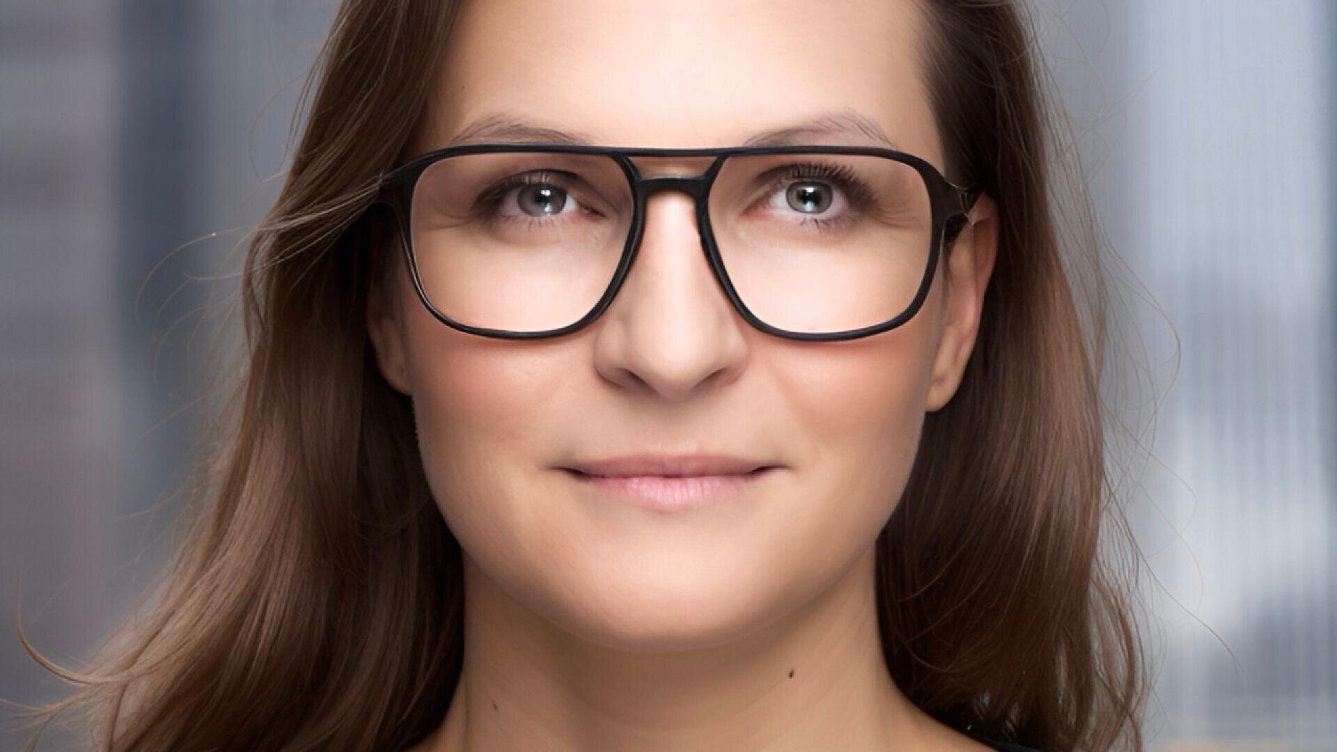 A woman with long brown hair and large black glasses looks straight ahead and smiles slightly, with a blurred background behind her.