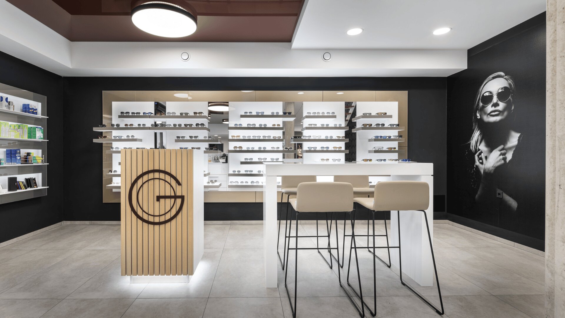 Modernes Brillengeschäft mit eleganten Brillenregalen, einem Holztresen mit einem großen "G"-Logo, hohen Hockern an einem weißen Tisch und einem schwarz-weißen Wandgemälde einer Frau mit Sonnenbrille an der Wand.