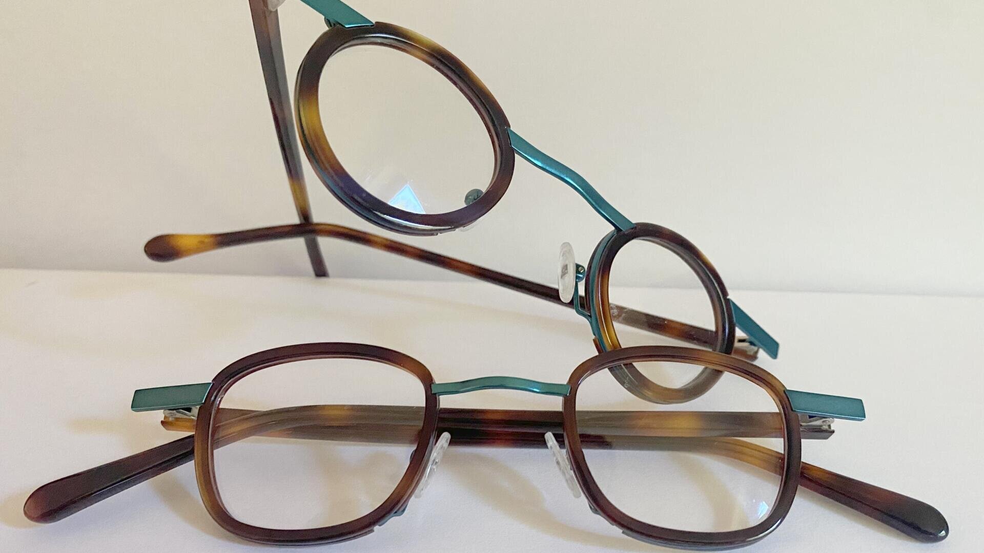 Zwei Brillen mit braunen Schildpattgestellen und blaugrünen Bügeln sind auf einer weißen Fläche angeordnet, wobei eine Brille flach liegt und die andere dahinter aufgestützt ist und beide nach vorne zeigen.