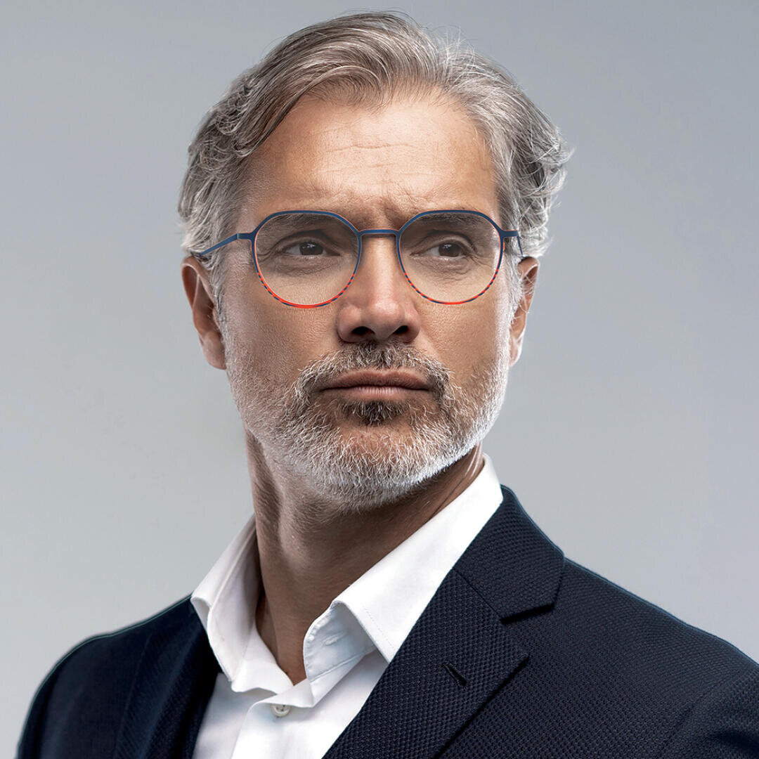Mann mittleren Alters mit grauem Haar und Bart, einer Brille mit blau-rotem Rand und einem dunklen Anzug über einem weißen Hemd, der vor einem schlichten Hintergrund selbstbewusst zur Seite blickt.