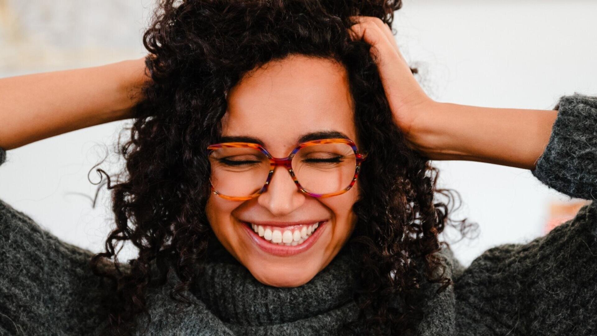 A woman with curly hair and colorful glasses smiles joyfully with her eyes closed, holding her hands in her hair. She is wearing a gray turtleneck sweater and appears happy and relaxed.