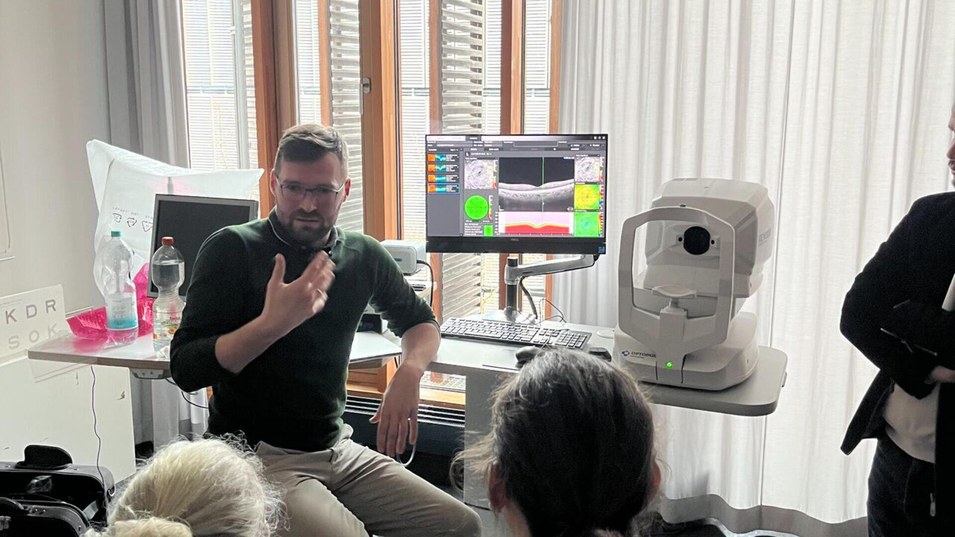 Ein Mann sitzt an einem Schreibtisch neben einem Augenscanner und einem Computer, der Augenscan-Bilder anzeigt, und gestikuliert, während er zu einer kleinen Gruppe von Menschen in einem hellen, modernen Raum spricht.
