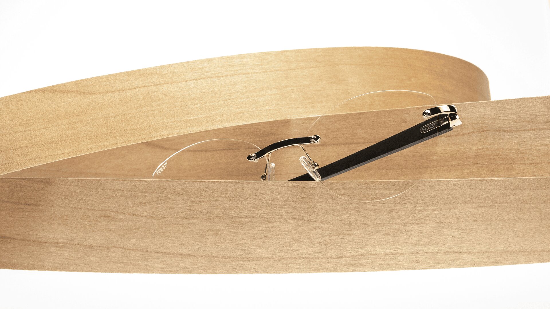 Eine minimalistische, randlose Brille mit schwarzen und goldenen Bügeln, die auf einem gebogenen, dünnen Streifen aus hellem Holz vor einem schlichten weißen Hintergrund ruhen und teilweise von diesem umhüllt sind.