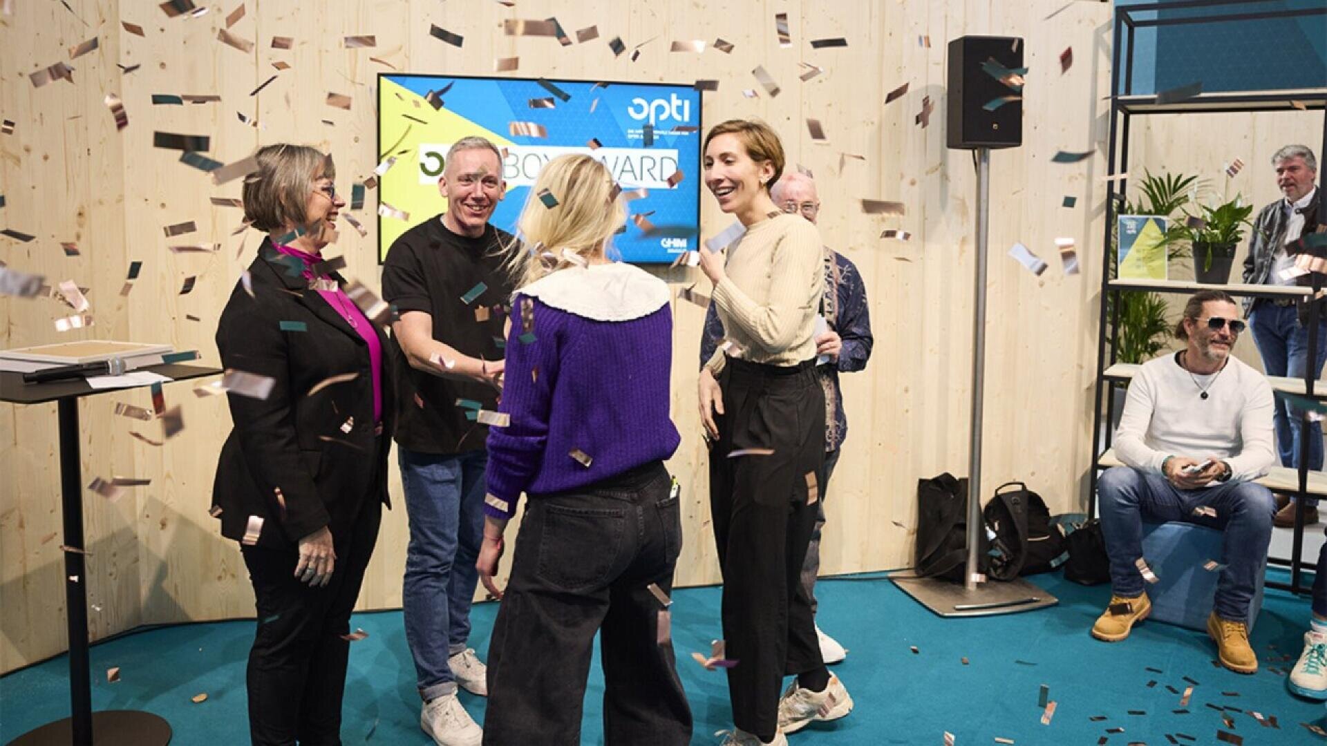 A group of people celebrate together indoors as confetti falls around them. Some are smiling and shaking hands, while others watch and clap. A screen and seated audience members are visible in the background.