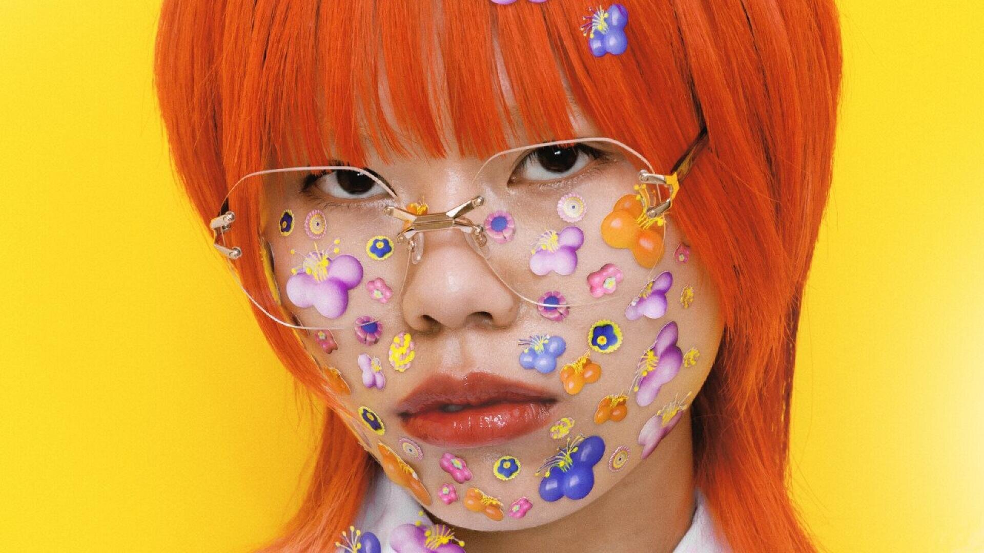A person with bright orange hair and large, clear glasses has their face covered with colorful flower and butterfly stickers, set against a vibrant yellow background.