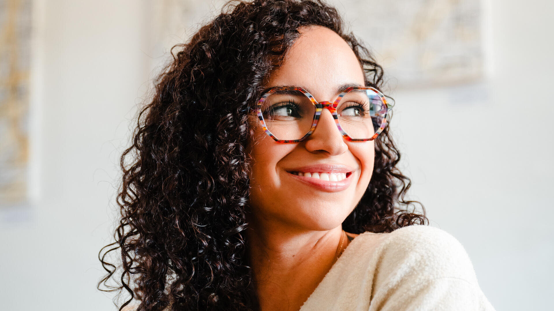 Eine Frau mit lockigem Haar und bunter, runder Brille lächelt und blickt zur Seite. Sie trägt ein helles Oberteil und befindet sich in einem Raum mit unscharfem Wandbild im Hintergrund.